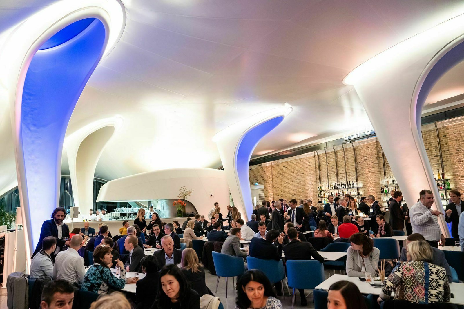 A modern, bustling restaurant with futuristic white and blue lighting features. Patrons sit at tables engaged in conversations against a backdrop of a lit-up bar and stylish decor. The ceiling curves elegantly, enhancing the dynamic atmosphere