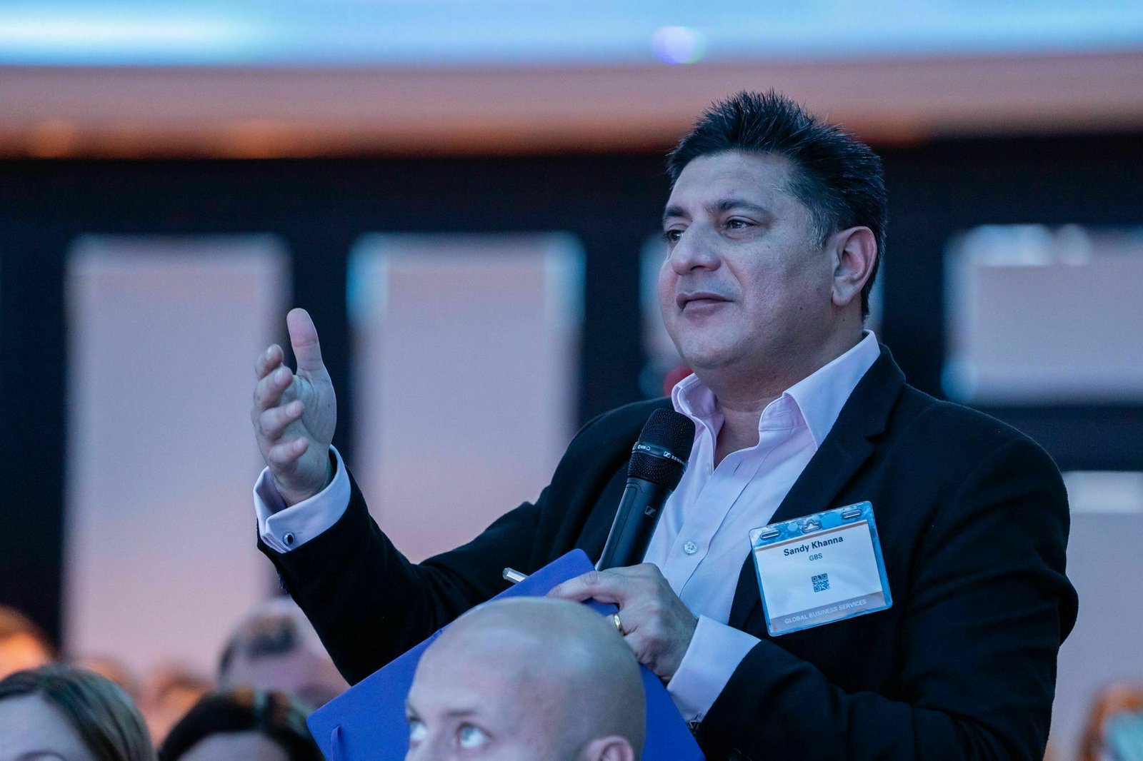 A man in a suit speaks into a microphone at an event. He gestures with his left hand, holding a folder, and wears a name tag. Several audience members are visible in the foreground. The background is softly blurred. James Gifford-Mead Photography - Event Photographer London