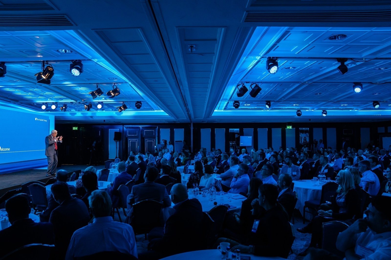 A speaker addresses a large audience in a dimly lit conference room. The room is bathed in blue light, with rows of seated attendees facing the stage. The ceiling has a grid pattern, and several round tables are arranged throughout the space
