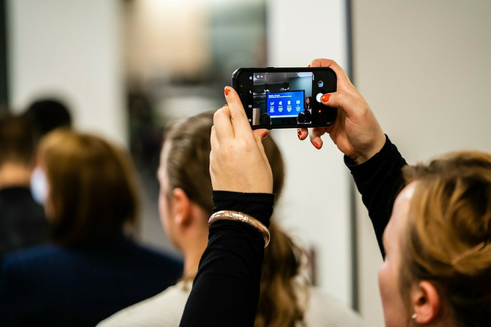A person with red nail polish holds up a smartphone to take a photo of a screen displaying a blue slide. The background features blurred individuals and a bright room. James Gifford-Mead Photography - Event Photographer London