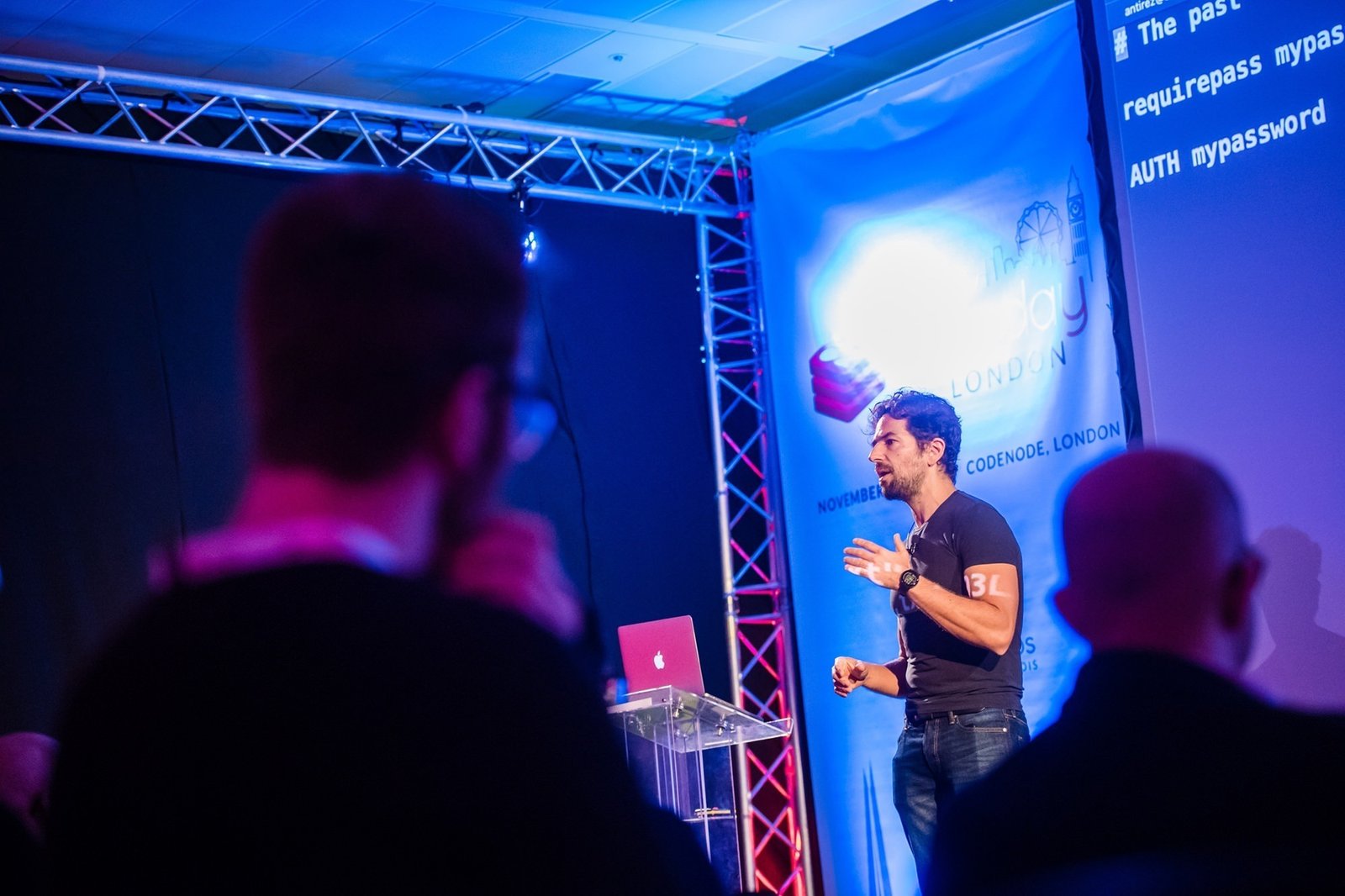 A man is giving a presentation on stage with a large screen displaying code. He is speaking to an audience in a dimly lit room. Two audience members are visible in the foreground. The setting appears to be a tech conference. James Gifford-Mead Photography - Event Photographer London