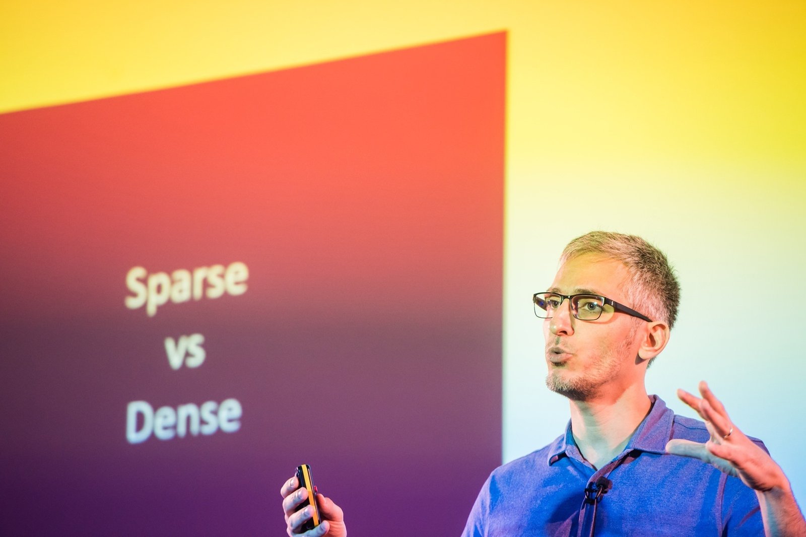 A person wearing glasses and a blue shirt speaks and gestures during a presentation. Behind them is a slide with the text "Sparse vs Dense" against a gradient background
