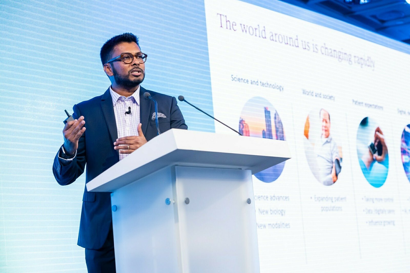 A person in a suit speaks at a podium with a microphone. Behind them is a large screen displaying a presentation slide titled "The world around us is changing rapidly" with related topics and images. James Gifford-Mead Photography - Event Photographer London