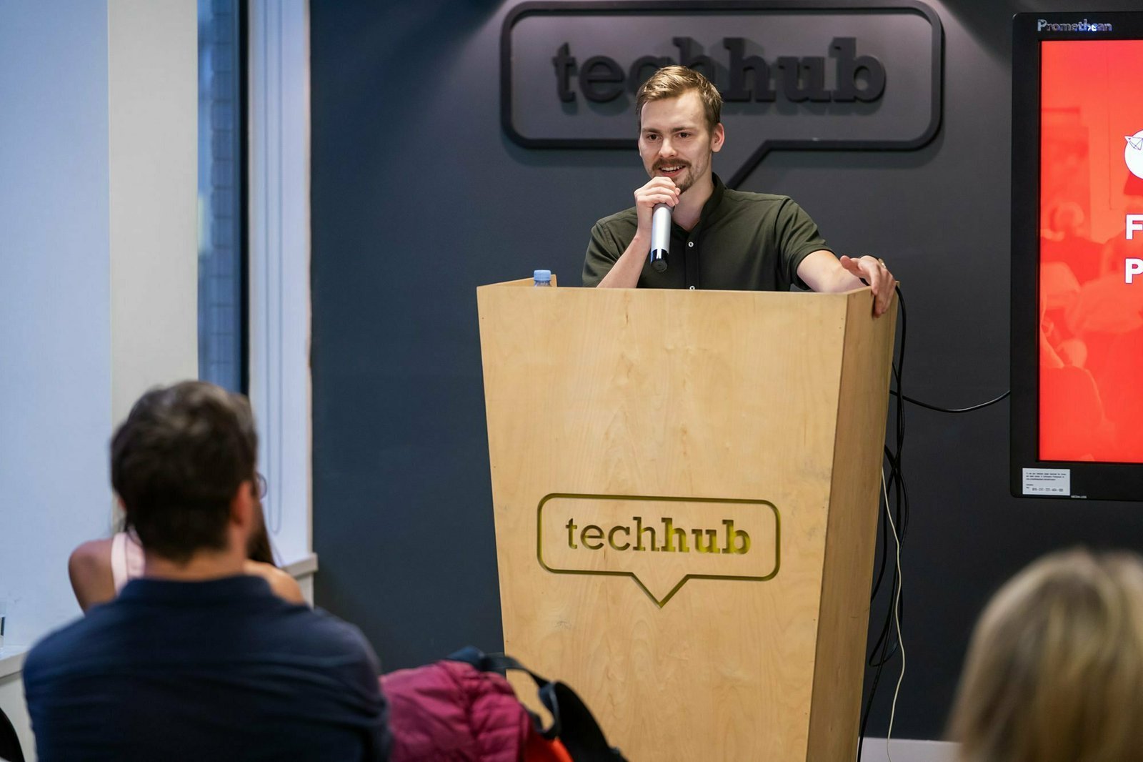 A person stands at a wooden podium with a "techhub" logo, speaking into a microphone. Several people are seated in the audience, and a red screen is partially visible to the side. James Gifford-Mead Photography - Event Photographer London