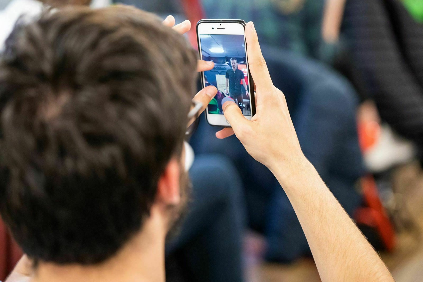 A person with short dark hair and glasses holds up a smartphone, taking a photo or video of someone or something in front. The phone screen displays a blurry image. The background is out of focus, with people and objects indistinctly visible. James Gifford-Mead Photography - Event Photographer London