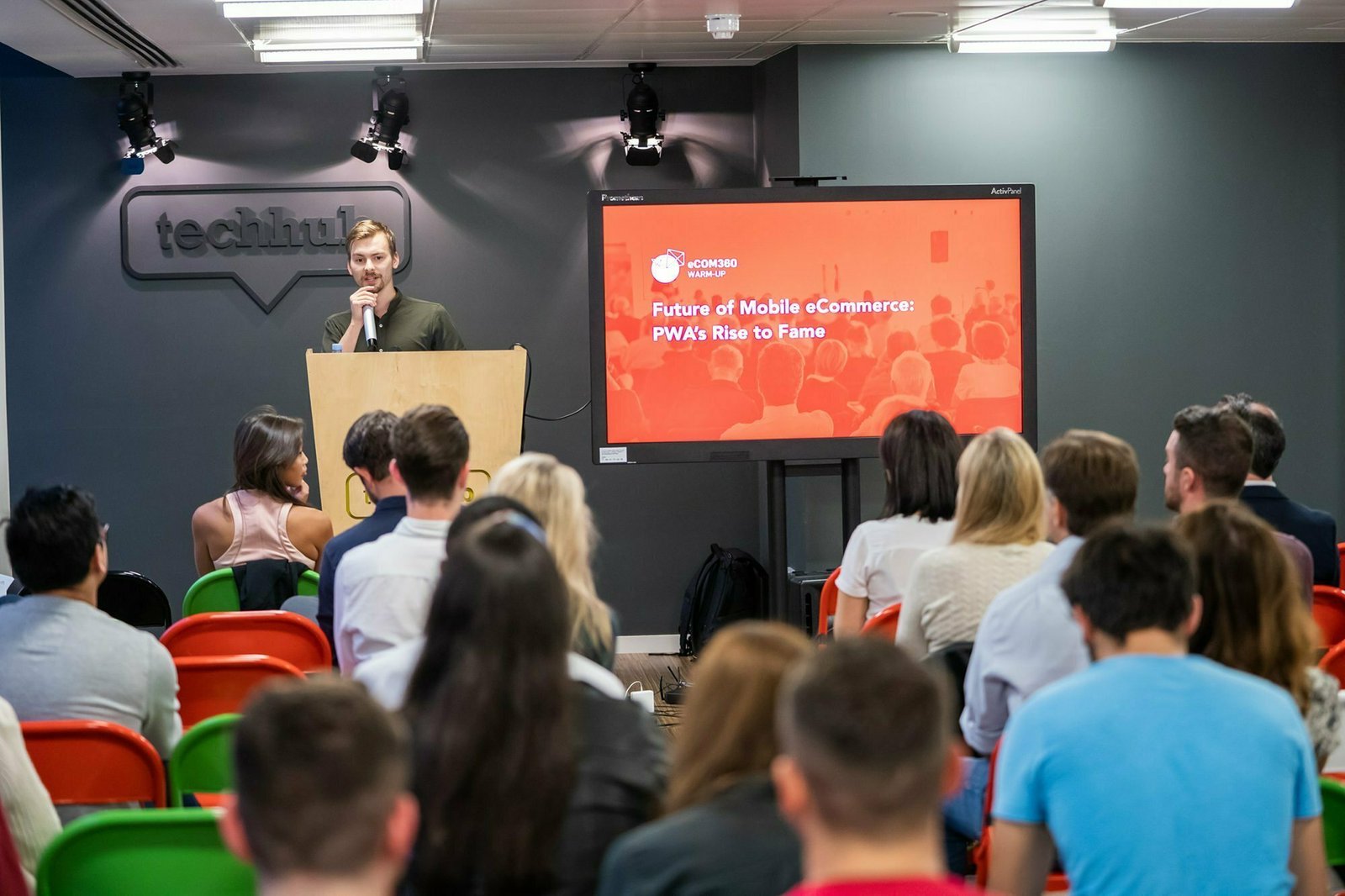 A person stands at a podium speaking to an audience seated in colorful chairs. Behind them, a screen displays a presentation titled "Future of Mobile eCommerce: PWA's Rise to Fame" in a modern conference room setting. James Gifford-Mead Photography - Event Photographer London