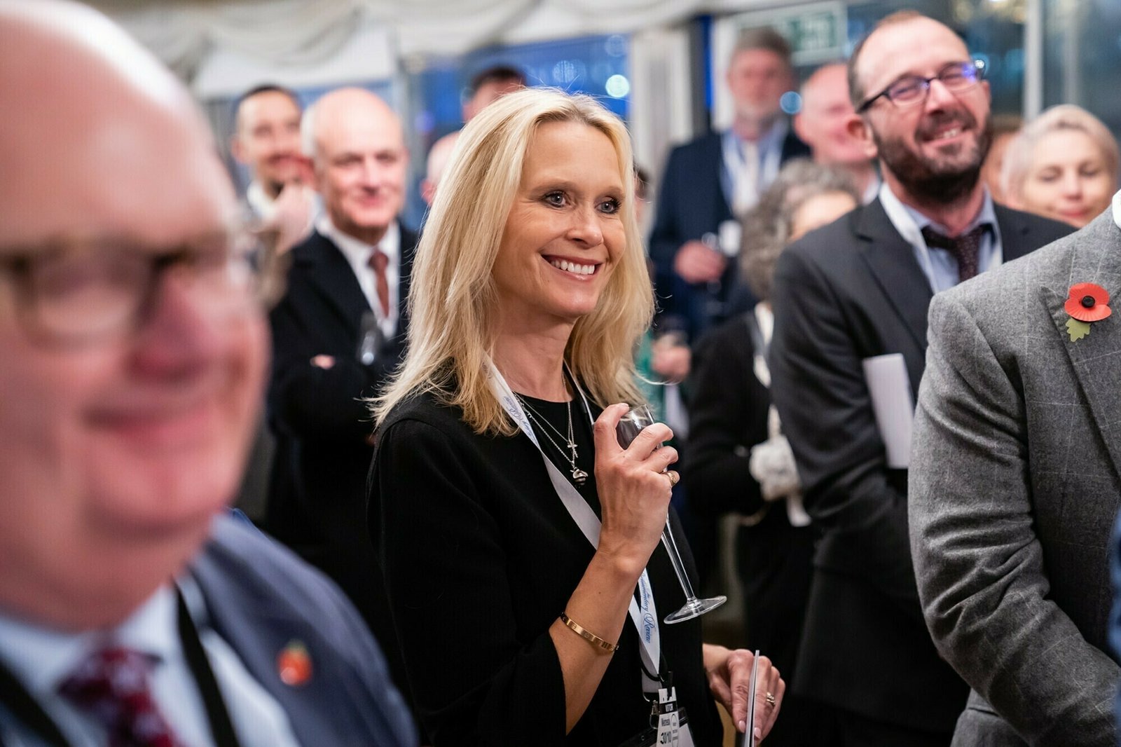 A group of people dressed in formal attire are gathered at an indoor event. A woman in the center is smiling and holding a wine glass. Others around her are also smiling and appear engaged. Some are wearing poppy pins. James Gifford-Mead Photography - Event Photographer London