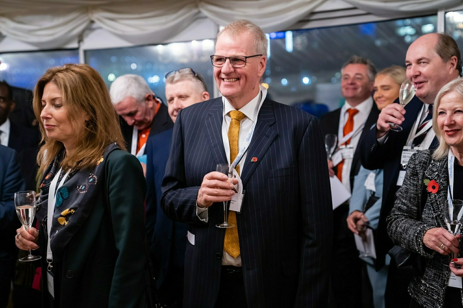 A group of people is standing together at an indoor event. They are dressed in formal attire and holding glasses, possibly for a toast. The background shows a lit-up interior with a large window. They appear to be smiling and engaged in conversation. James Gifford-Mead Photography - Event Photographer London