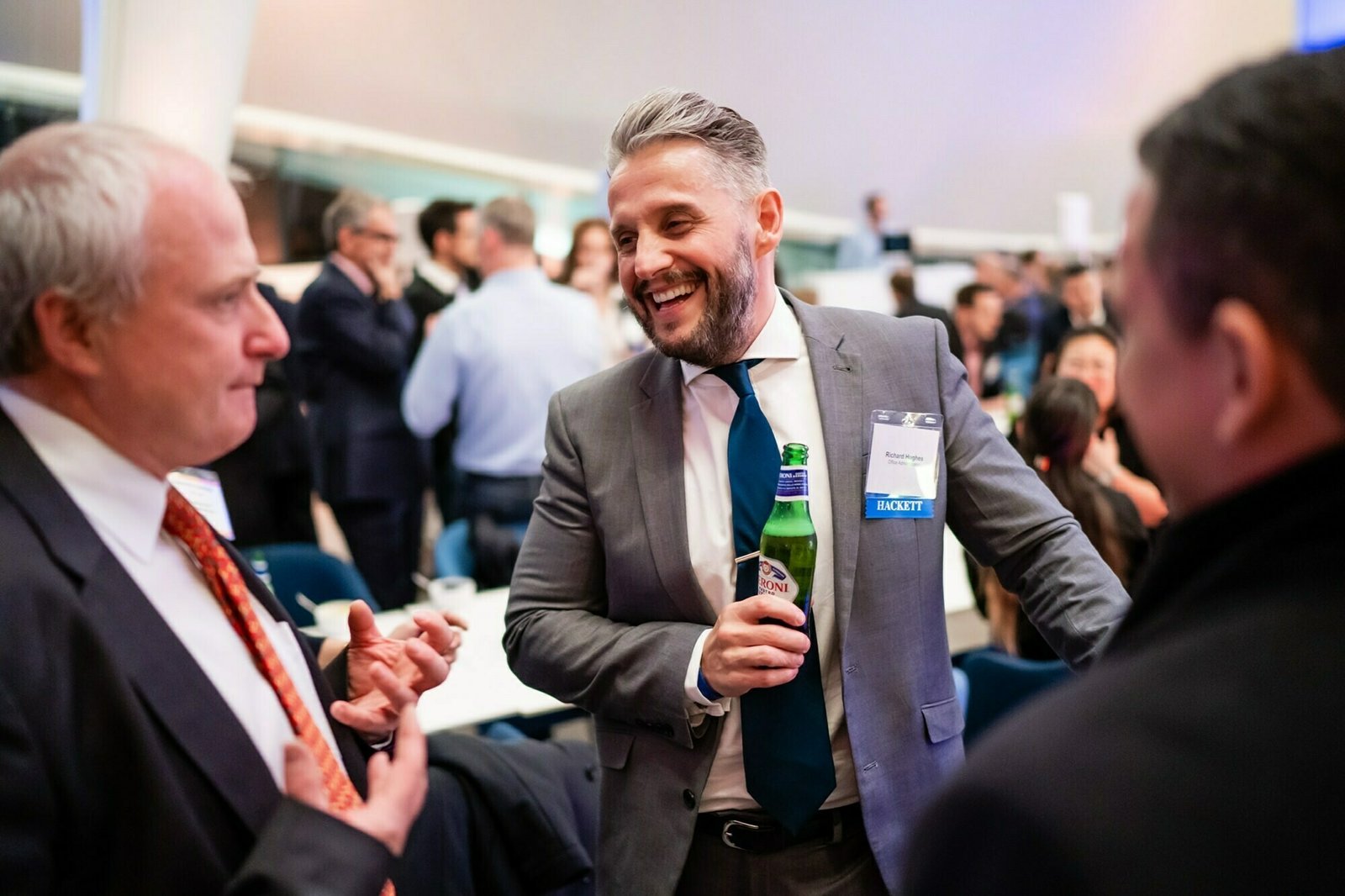 A man in a suit with a name badge laughs while holding a beer. Two other men, also in suits, are engaged in conversation with him. The setting appears to be a social or networking event with several people in the background. James Gifford-Mead Photography - Event Photographer London