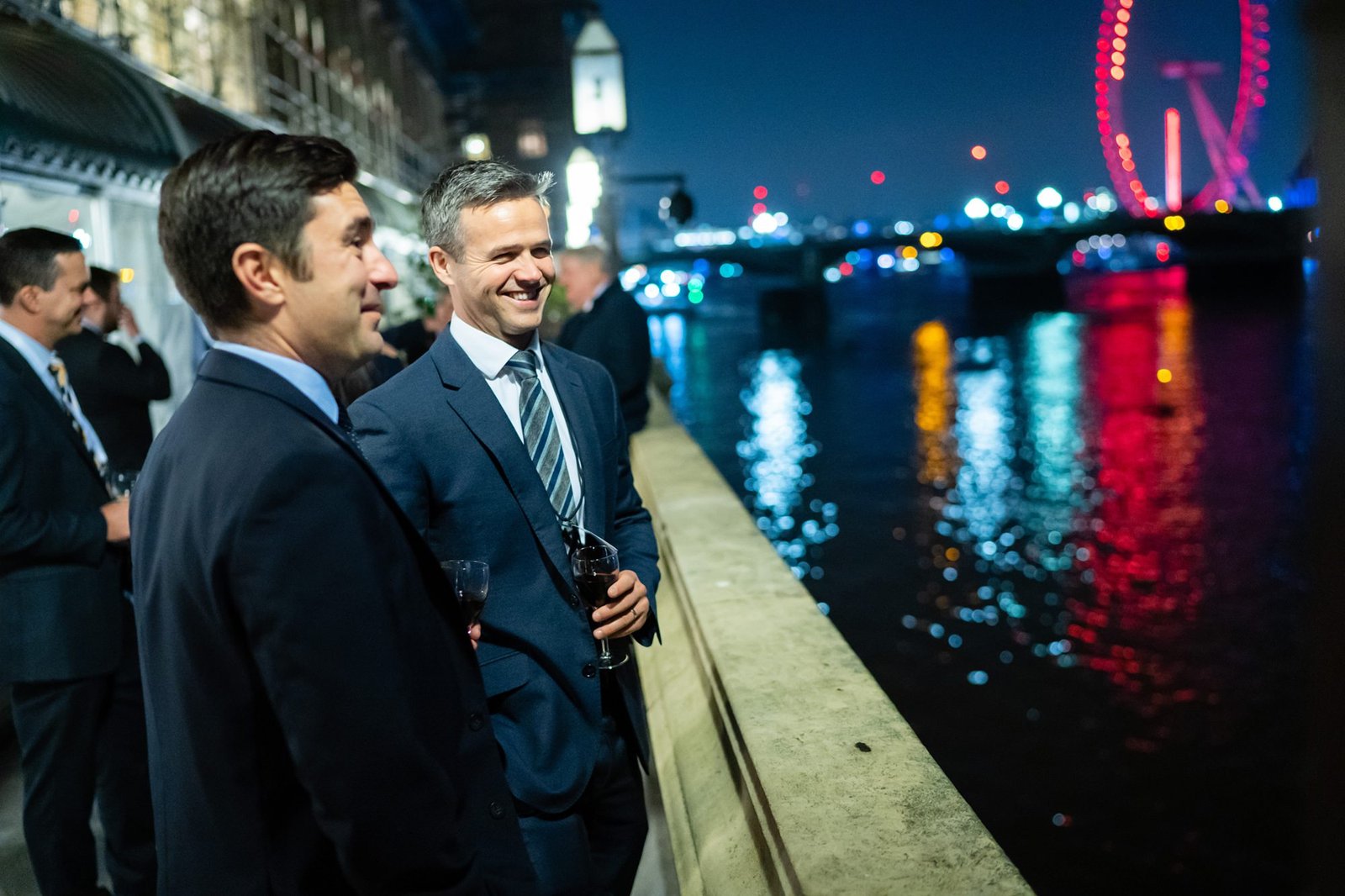 Two men in suits stand by a riverside at night, engaged in conversation. They hold drinks and smile. In the background, a cityscape with bright lights and a large Ferris wheel is visible reflecting on the water