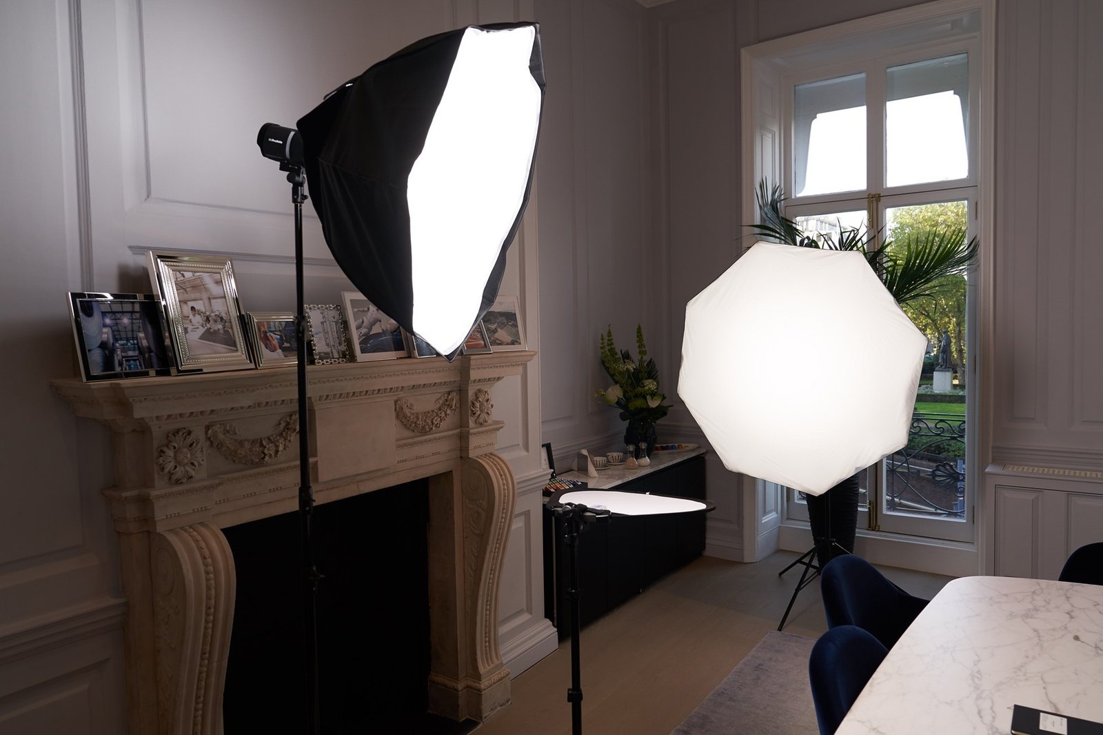 Photography lighting setup in a room with softboxes placed near a fireplace. Framed photos are on the mantel, and a large window offers a view of greenery outside. A table with chairs is partially visible in the foreground