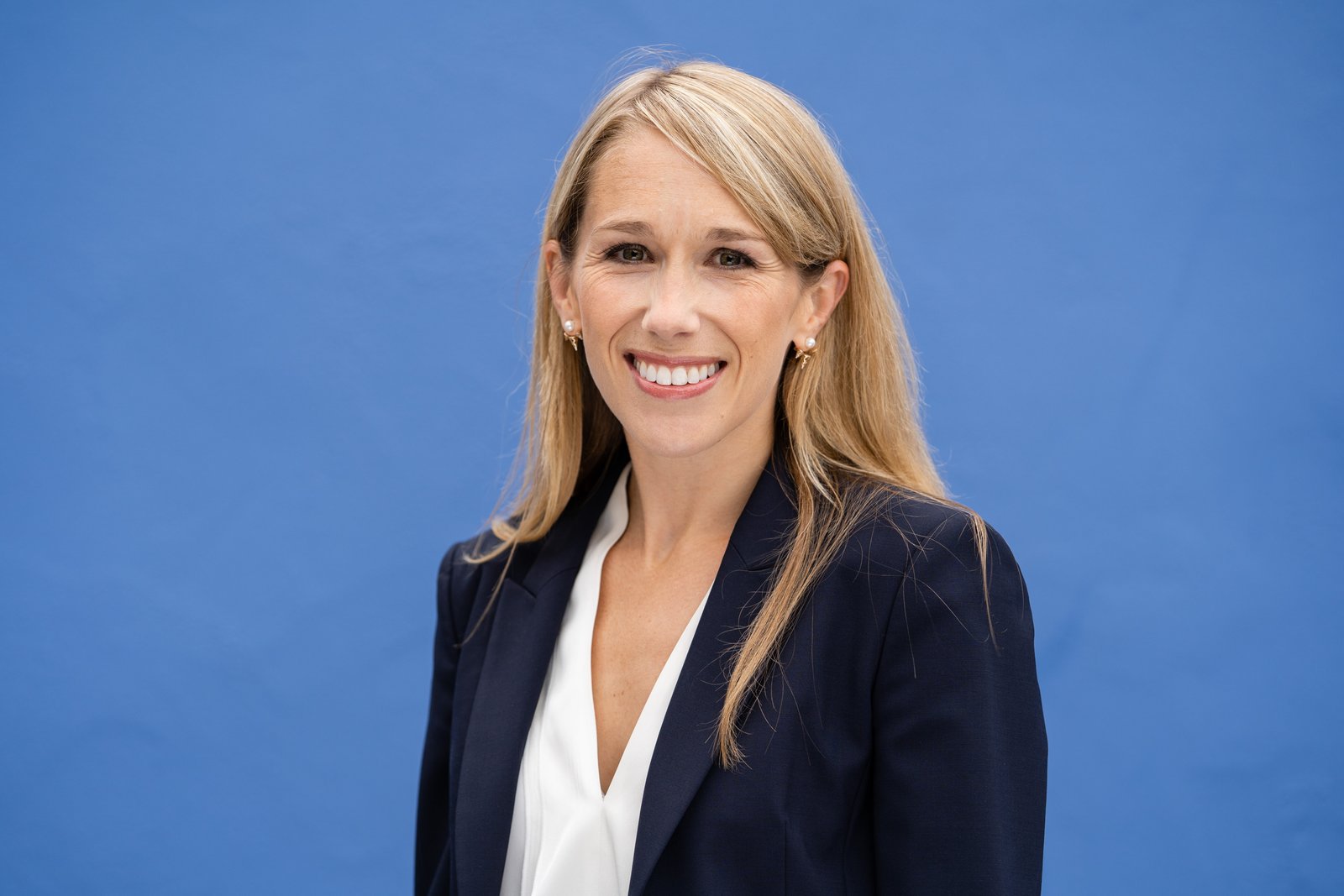A smiling woman with long blonde hair is wearing a navy blazer over a white top. She is standing against a plain blue background