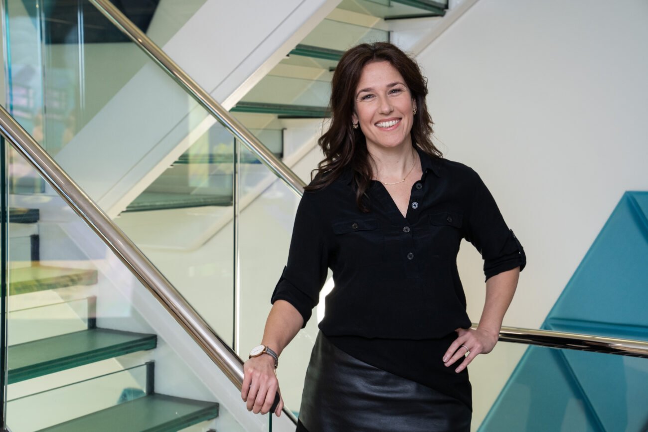 A person with long brown hair stands confidently on a modern glass staircase, wearing a black blouse and skirt. They have one hand on their hip and are smiling at the camera. James Gifford-Mead Photography - Event Photographer London