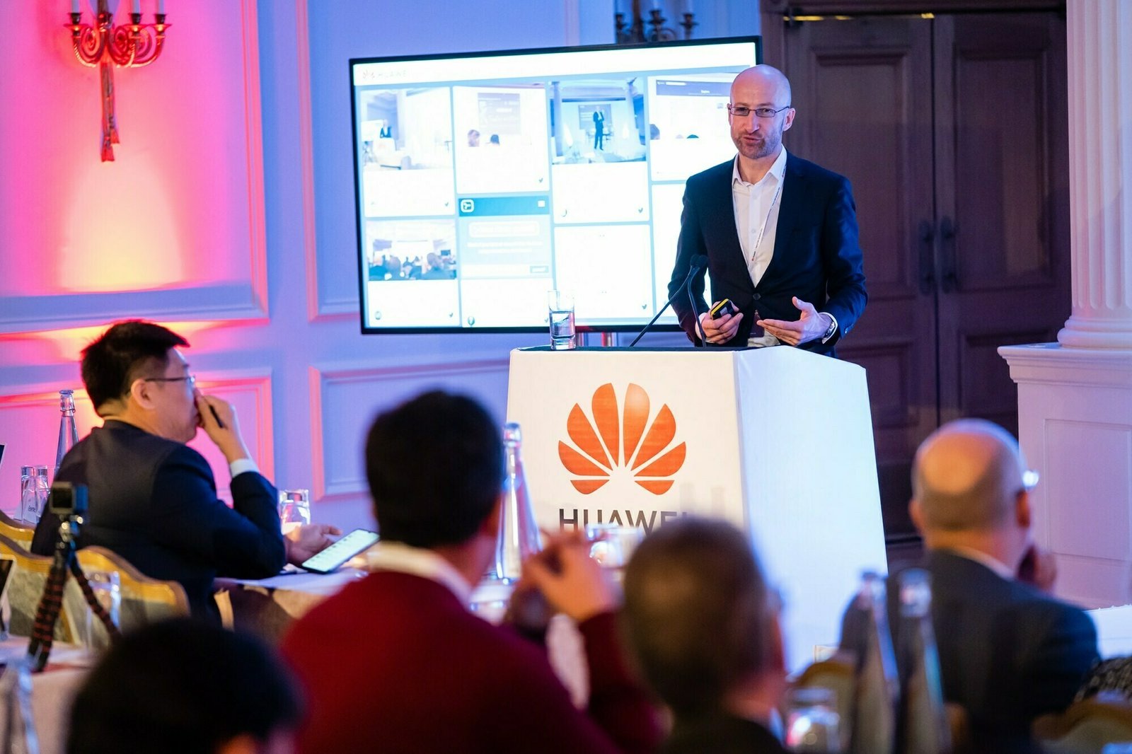 A man in a suit stands at a podium with a Huawei logo, giving a presentation to an audience. A large screen displays various images behind him. The room is decorated with red lighting and elegant white walls. James Gifford-Mead Photography - Event Photographer London