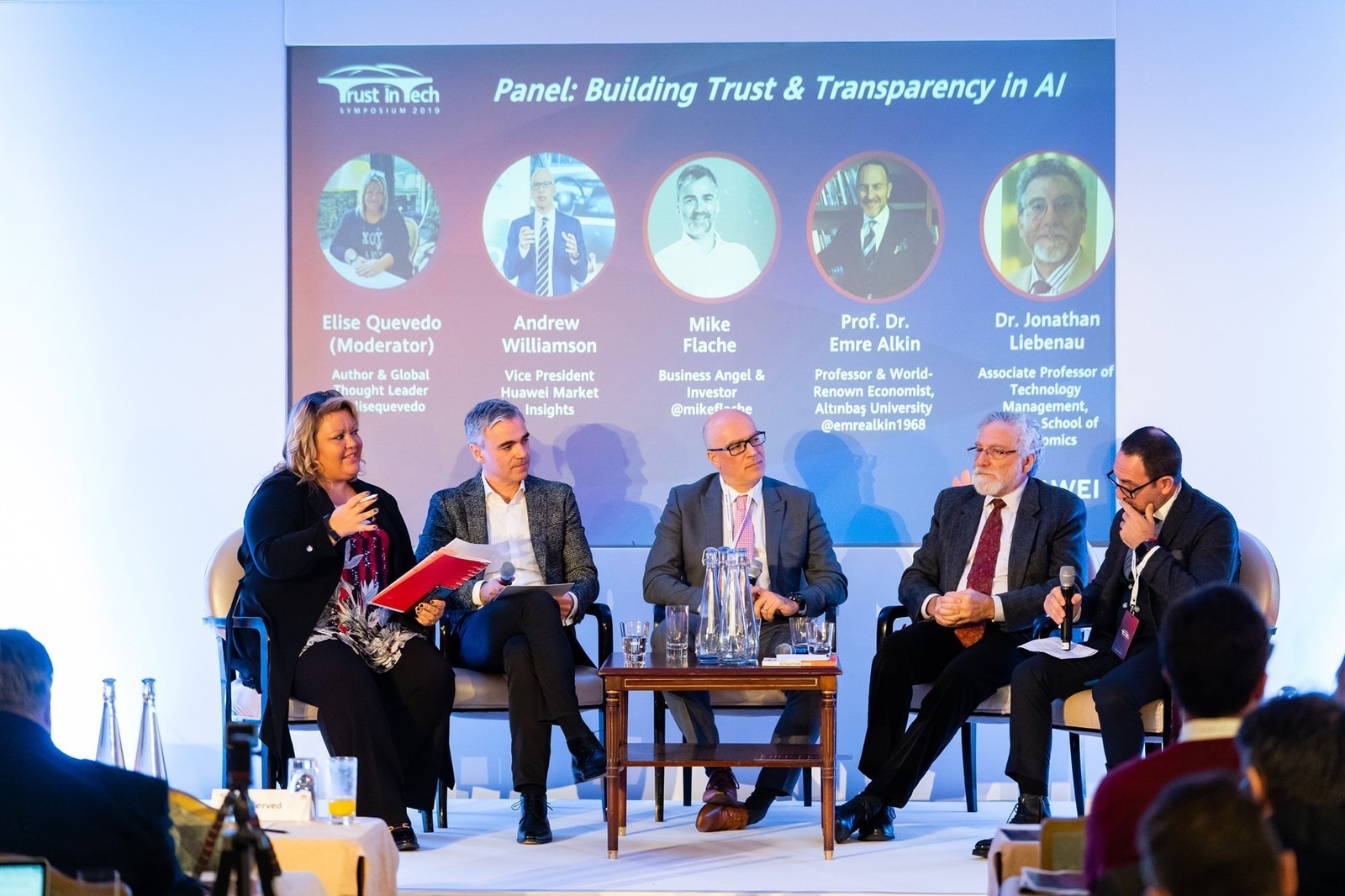 Five individuals are seated on a stage having a panel discussion titled "Building Trust & Transparency in AI." There is a screen in the background displaying their names and titles. The moderator is holding a red folder