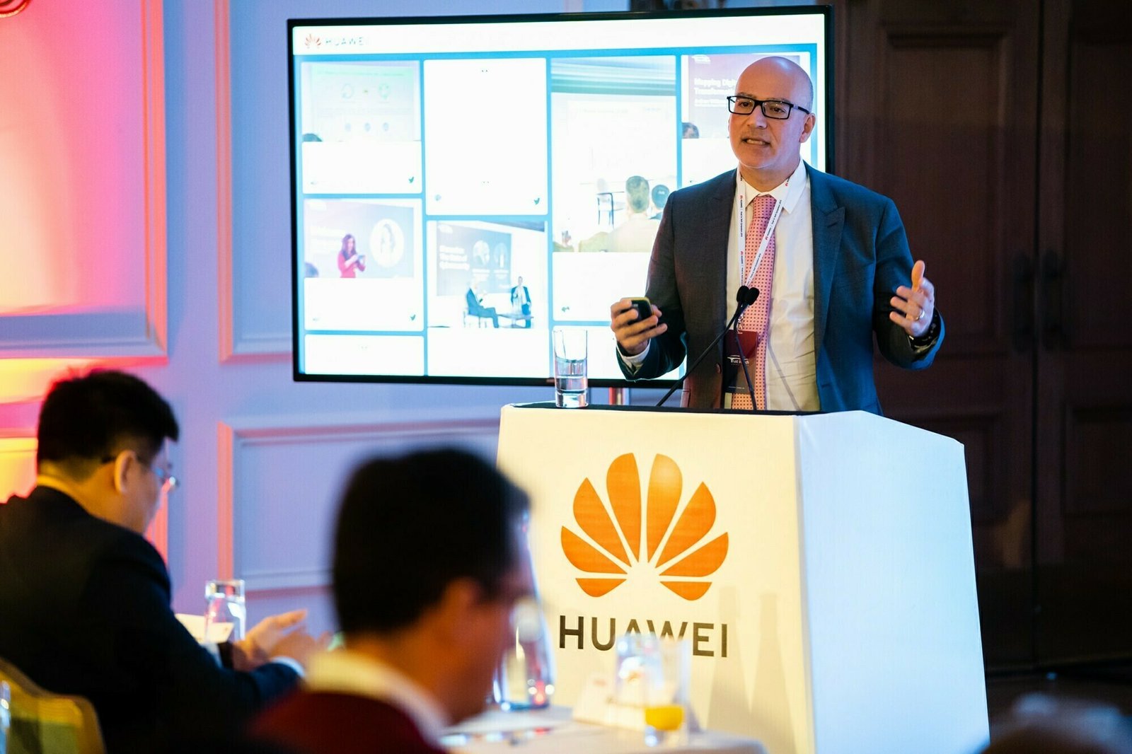 A man in a suit and glasses stands at a podium with the Huawei logo, speaking into microphones. Behind him is a screen displaying various slides. Seated attendees are visible in the foreground. James Gifford-Mead Photography - Event Photographer London
