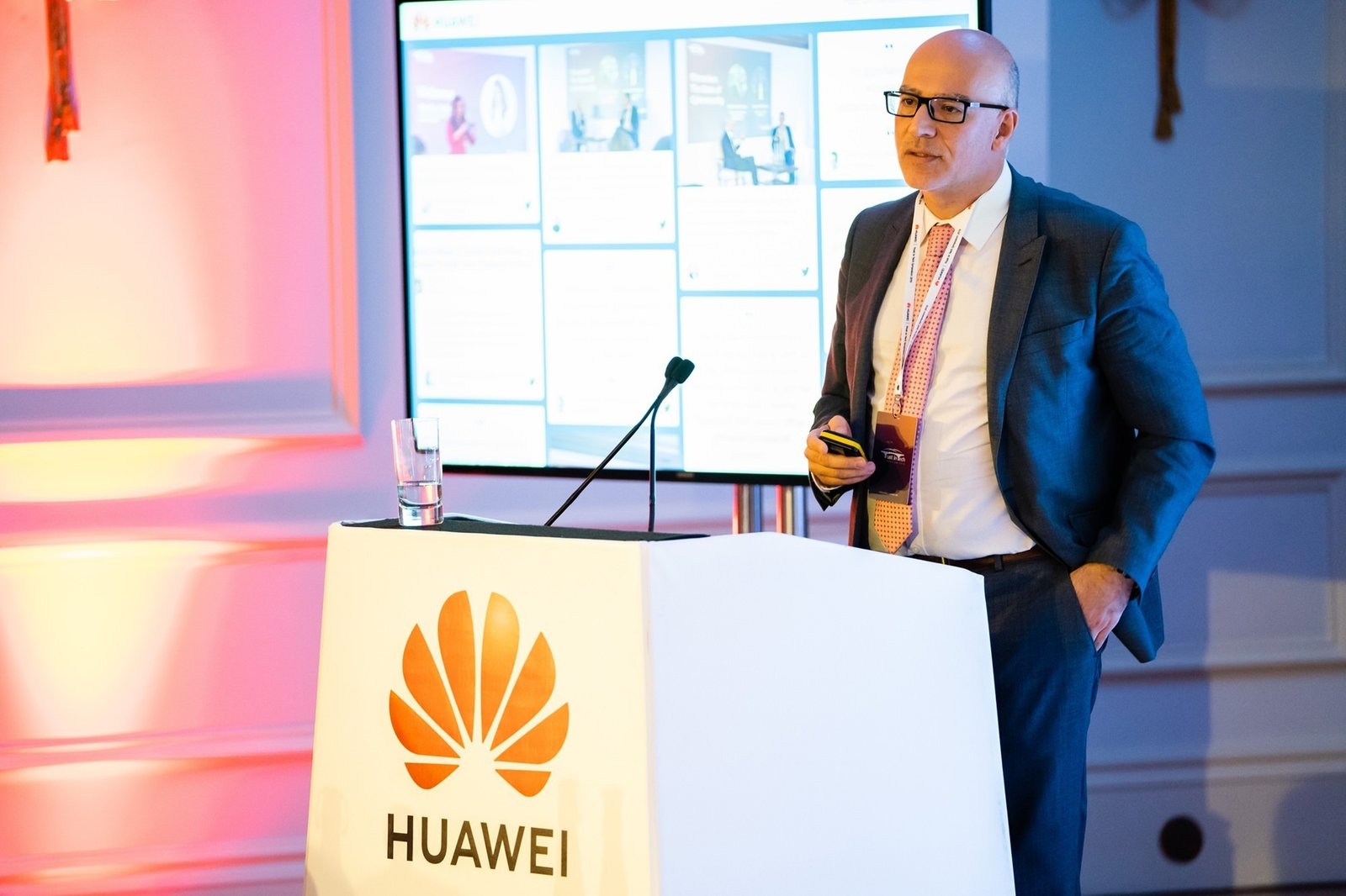 A man in a suit stands at a podium with the Huawei logo, speaking into a microphone. Behind him is a digital screen displaying slides. The room is lit with a soft orange and blue light. James Gifford-Mead Photography - Event Photographer London
