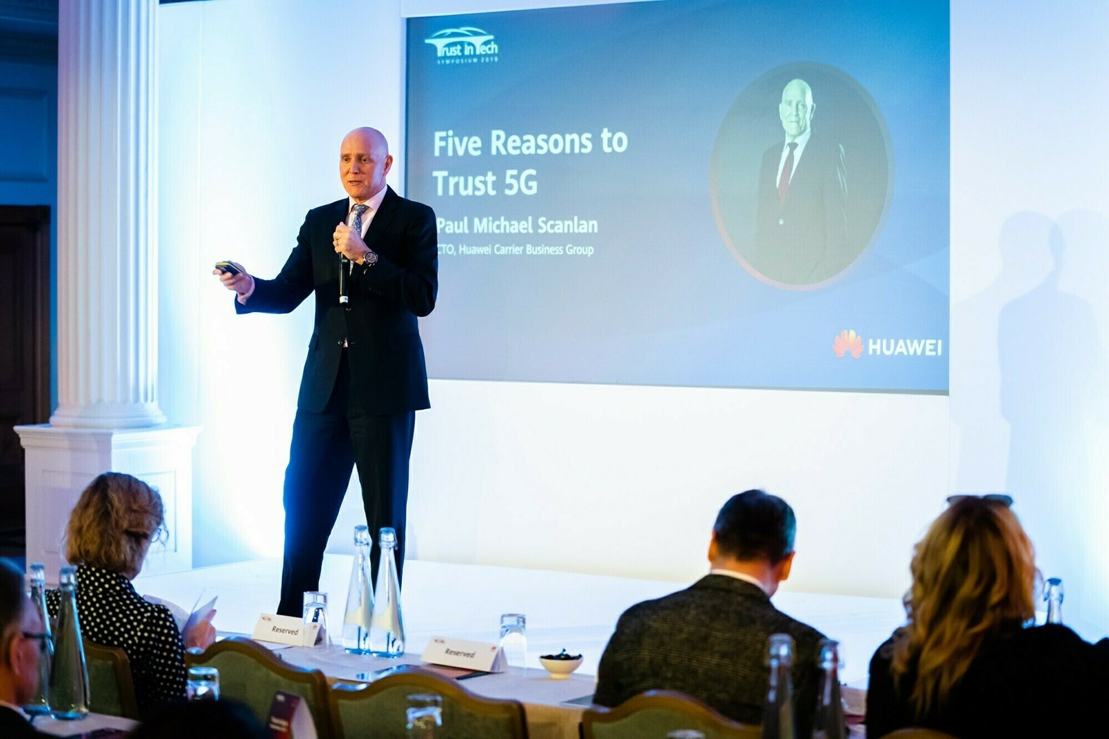 A man in a suit is giving a presentation titled "Five Reasons to Trust 5G" on stage. A slide with a photo and text accompanies him. The audience is seated in rows, taking notes. Bottles and notepads are on the tables. James Gifford-Mead Photography - Event Photographer London