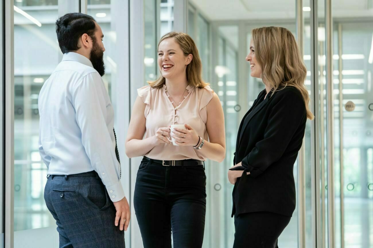 Three people are standing in an office hallway, smiling and engaged in a conversation. One person holds a cup, while the others gesture with their hands. Captured by a corporate photographer in London, they appear relaxed and professional, dressed in business attire. James Gifford-Mead Photography - Event Photographer London