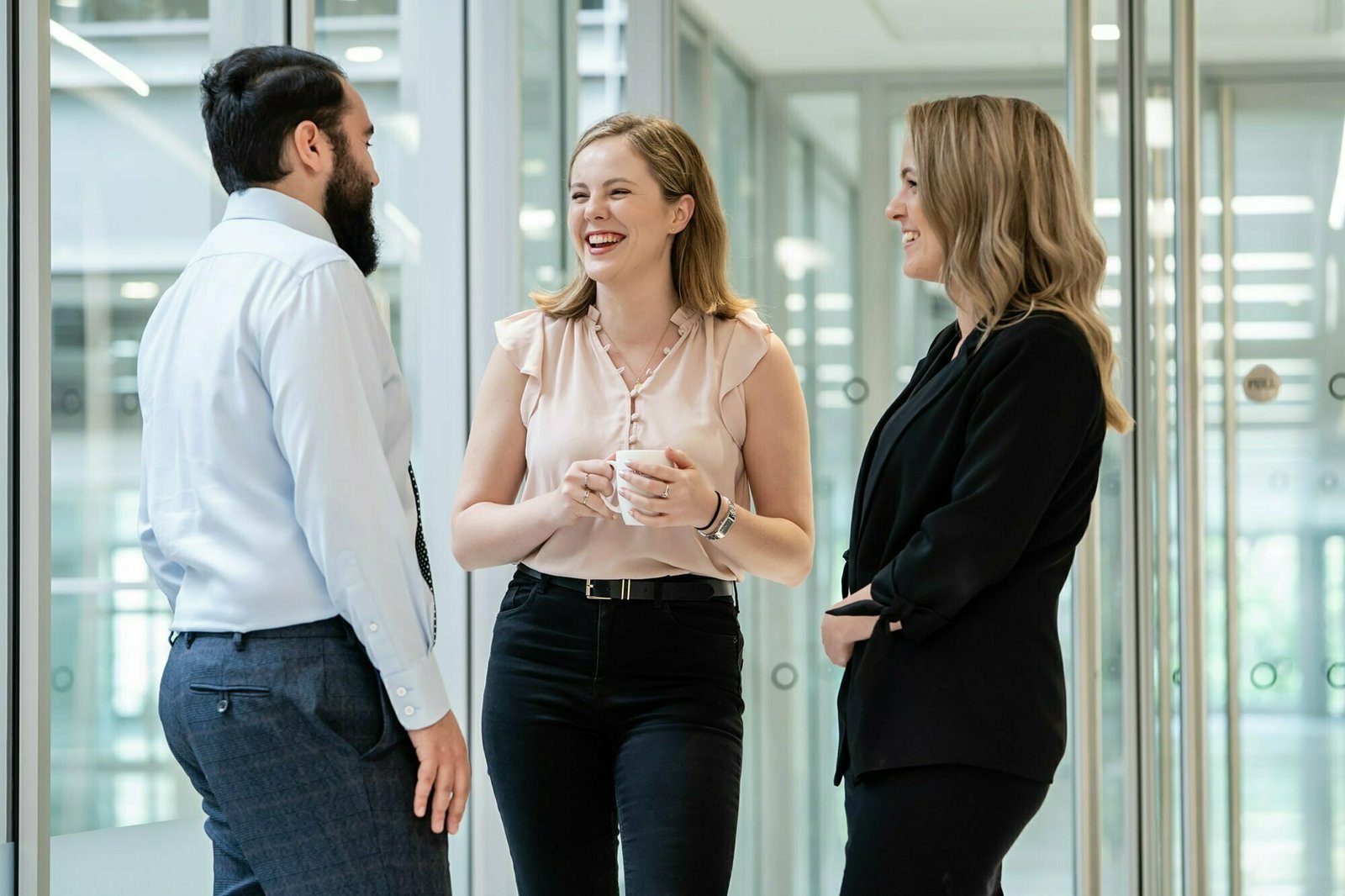 Three people are standing in an office hallway, smiling and engaged in a conversation. One person holds a cup, while the others gesture with their hands. Captured by a corporate photographer in London, they appear relaxed and professional, dressed in business attire. James Gifford-Mead Photography - Event Photographer London