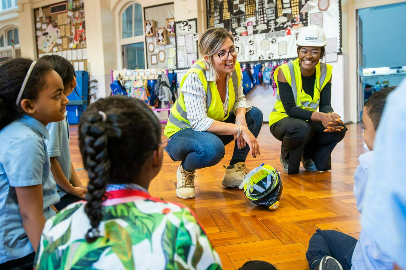 Two women in safety vests and helmets engage with young students sitting in a circle on a wooden floor. Captured by a talented corporate photographer in London, the vibrant classroom scene features colorful artwork on the walls. The women are smiling, with one sporting a hard hat