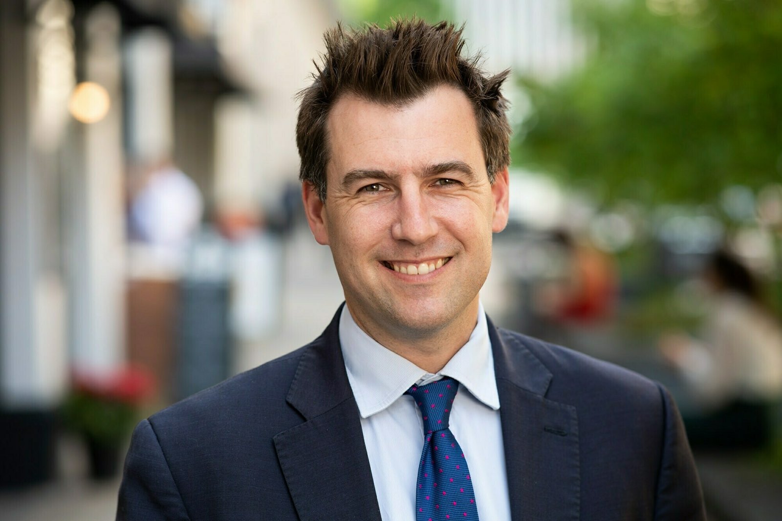 A man with short, spiky hair wearing a dark suit, light blue shirt, and blue tie with pink dots, smiling outdoors. The background is an urban street with blurred greenery and people