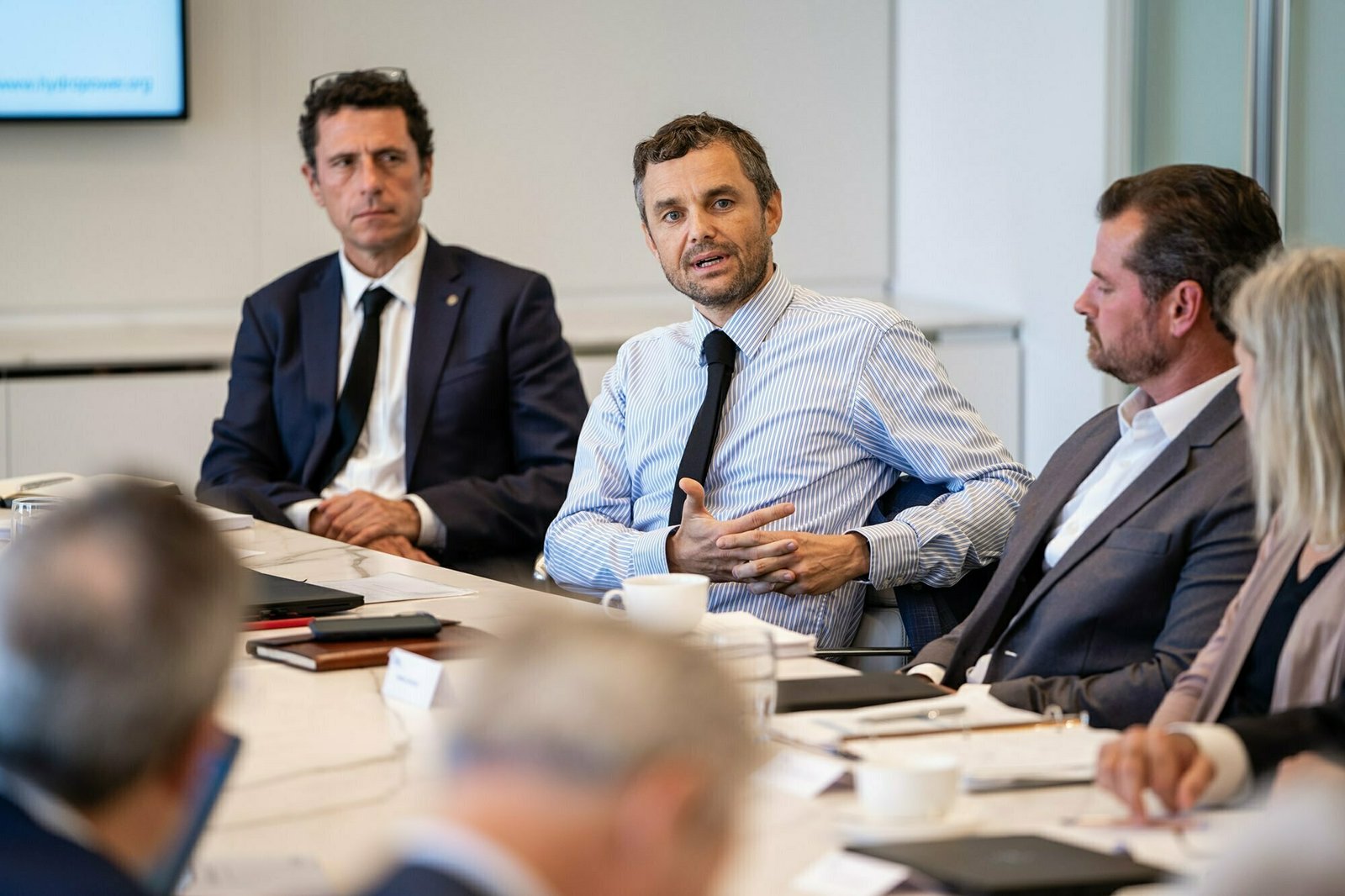 A group of people in business attire are seated around a conference table. A man in a striped shirt and tie is speaking, while others listen. Papers, notebooks, and cups are on the table in a modern office setting. James Gifford-Mead Photography - Event Photographer London