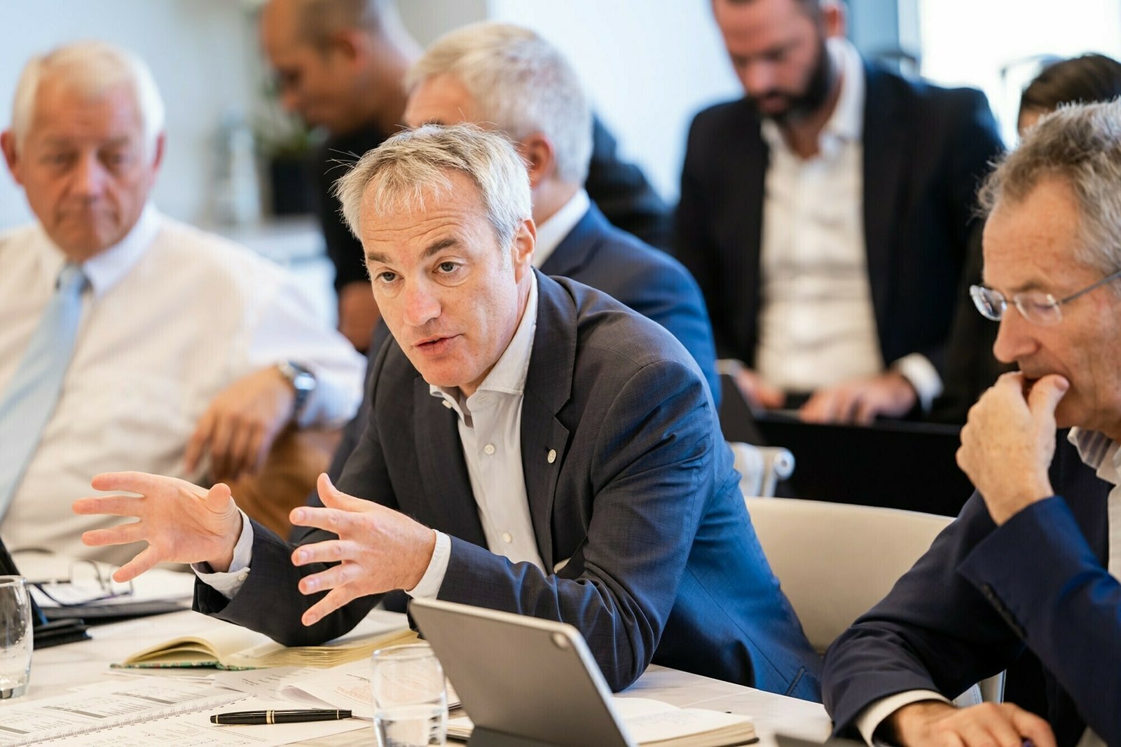 A group of people in business attire are engaged in a meeting around a table. One man in the center is speaking, gesturing with his hands. Documents and a tablet are on the table, and others are listening attentively. James Gifford-Mead Photography - Event Photographer London