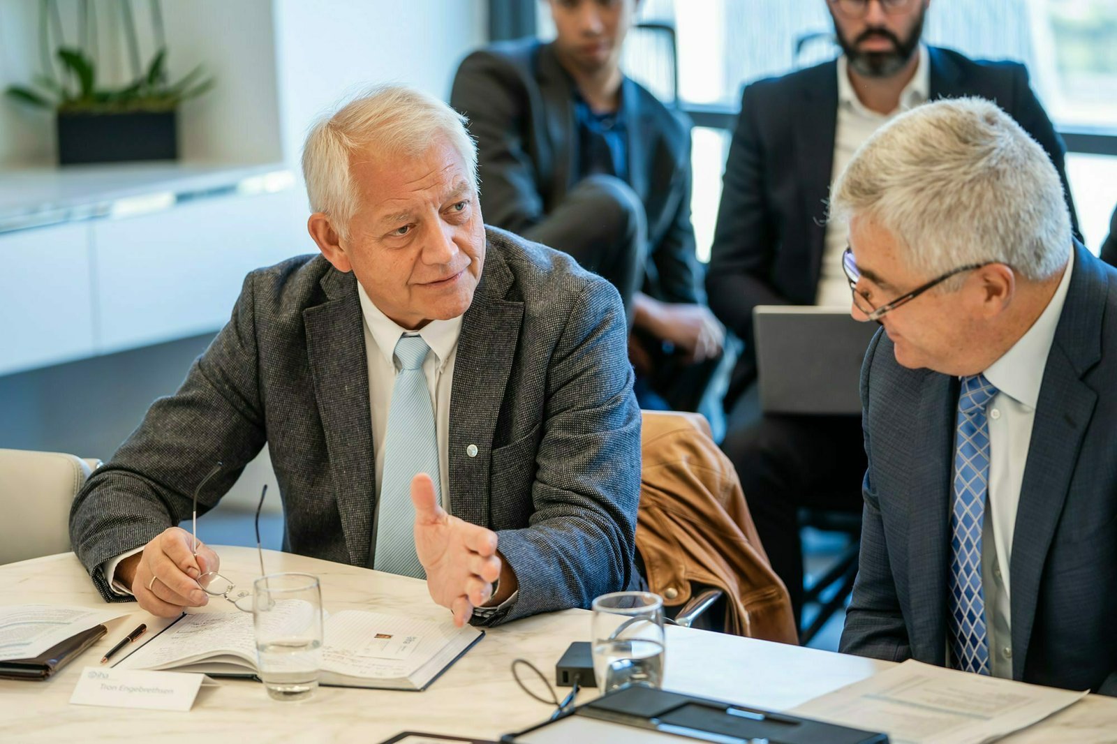 Two elderly men in suits are engaged in conversation at a conference table. Both have gray hair and are surrounded by papers and glasses of water. Other individuals sit in the background, some using laptops. James Gifford-Mead Photography - Event Photographer London