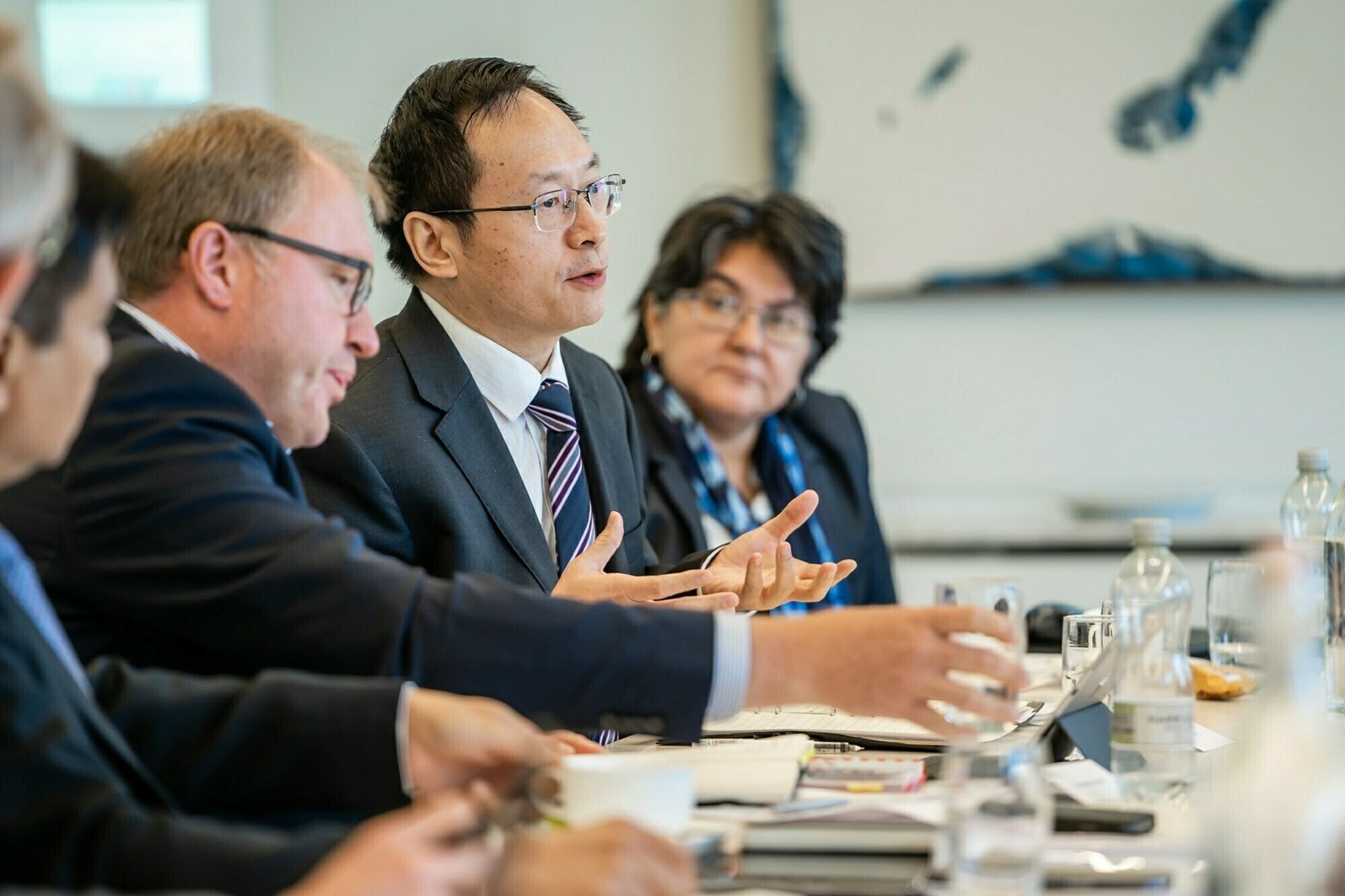 A group of business professionals in formal attire are seated at a conference table engaged in discussion. The focus is on a man speaking, gesturing with his hands. Papers, notebooks, and bottles are on the table. A blurred painting is in the background. James Gifford-Mead Photography - Event Photographer London