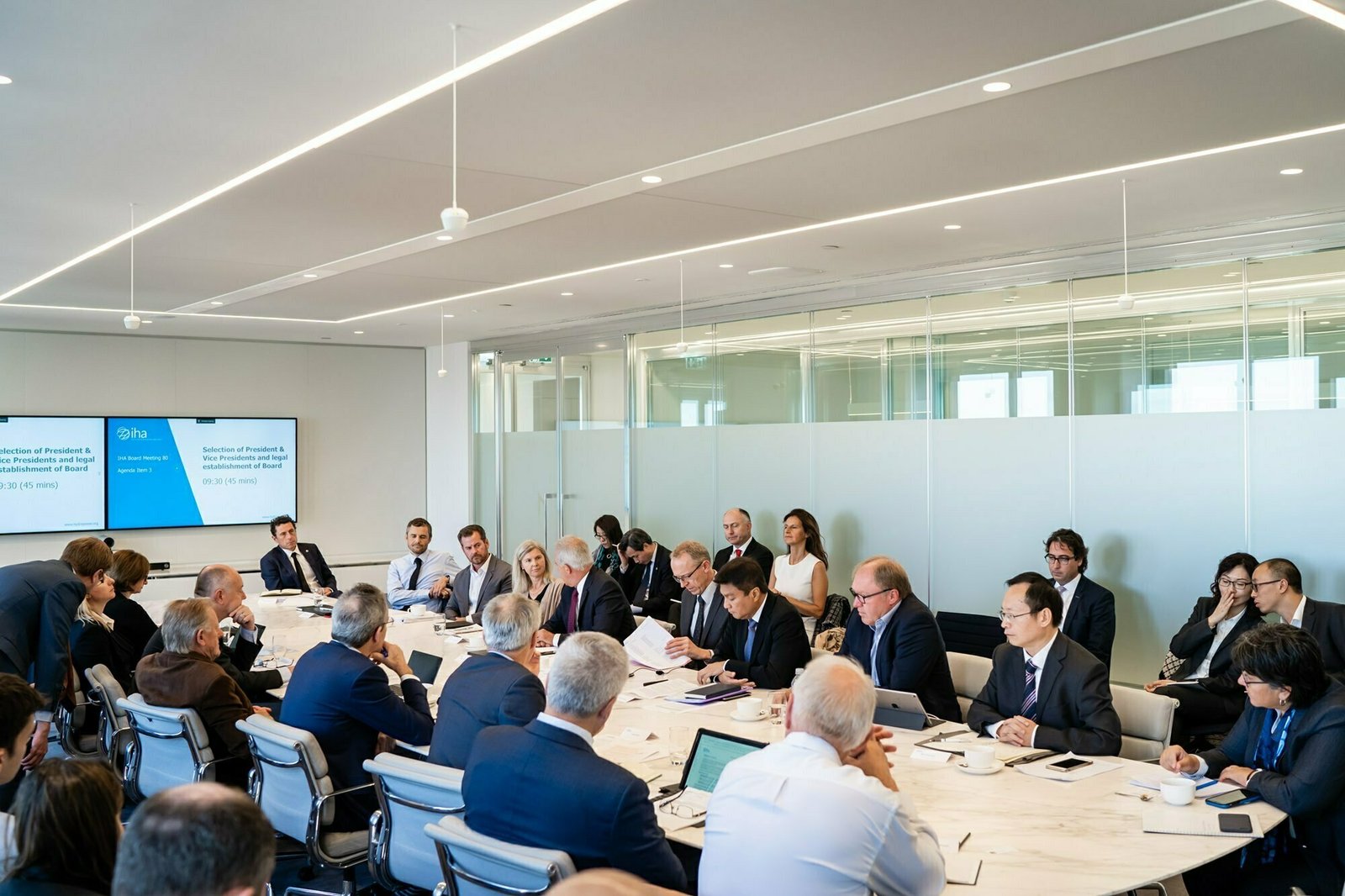 A large group of people in business attire sit around a U-shaped conference table in a modern meeting room. Two screens display a presentation. The room has white walls, glass partitions, and ceiling lights