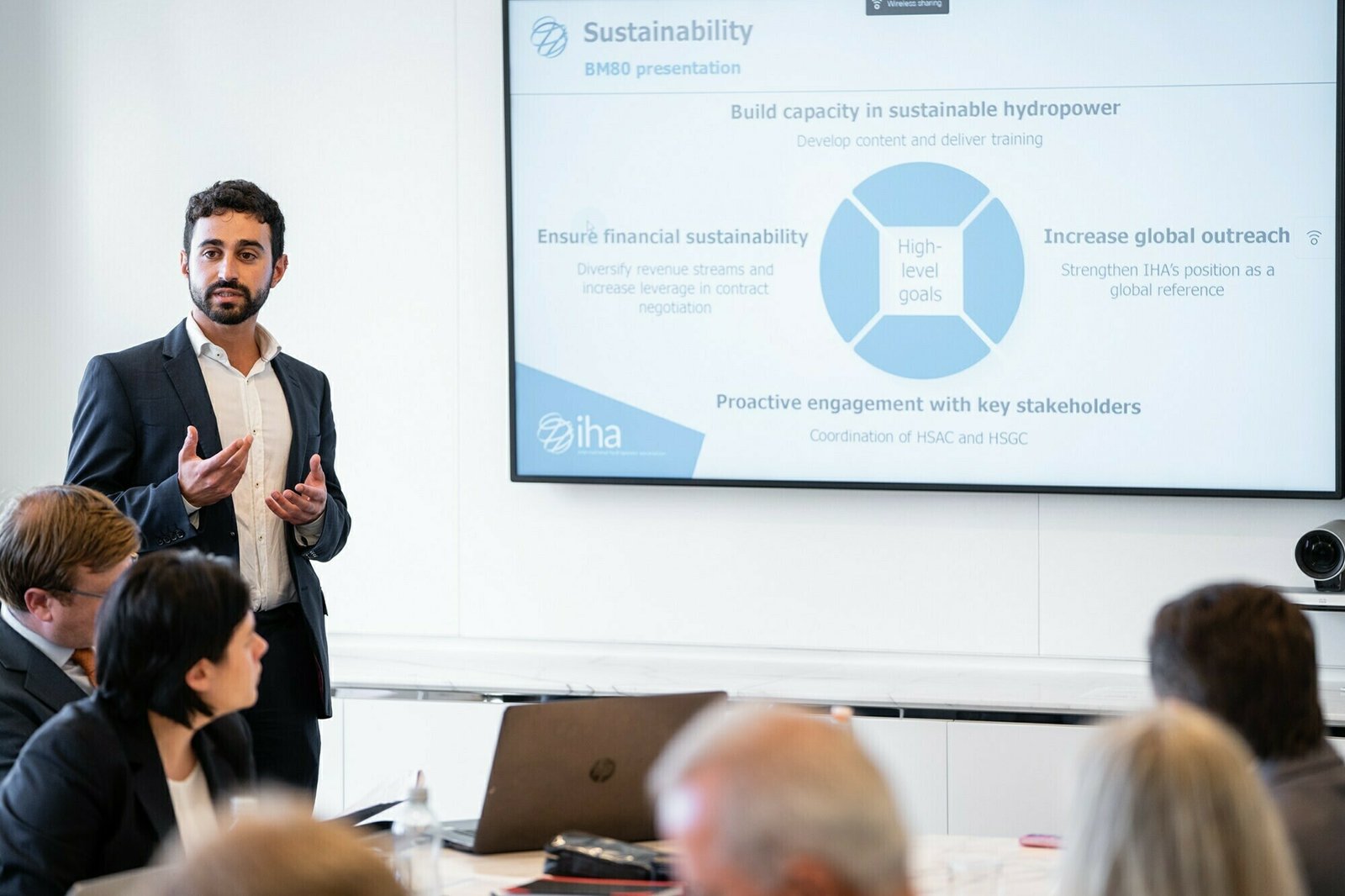 A man presents in a conference room with a screen displaying a slide on sustainability in hydropower. Seated attendees listen attentively. Key points include financial sustainability, global outreach, engagement, and goals. James Gifford-Mead Photography - Event Photographer London