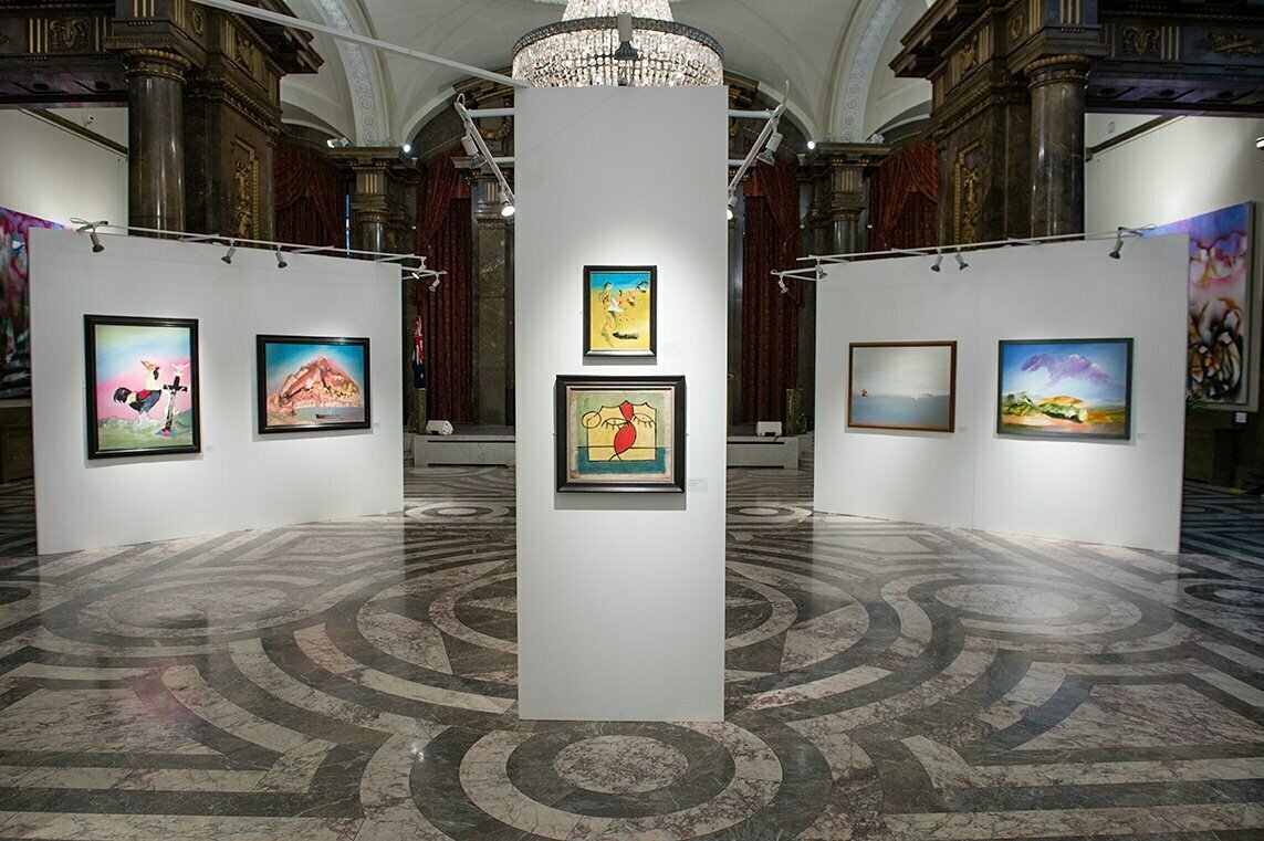 The art gallery interior, perfect for a corporate photographer in London, boasts a central white wall with vibrant paintings. Ornate columns, marble-patterned flooring, and a chandelier add elegance, while additional artworks adorn the surrounding white walls