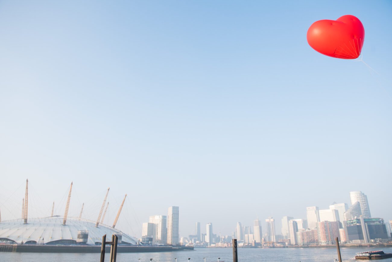 A red heart-shaped balloon floats in the clear sky above a city skyline. In the foreground, captured by a corporate photographer in London, a dome-shaped building with a distinctive roof sits by the water, framed by modern skyscrapers