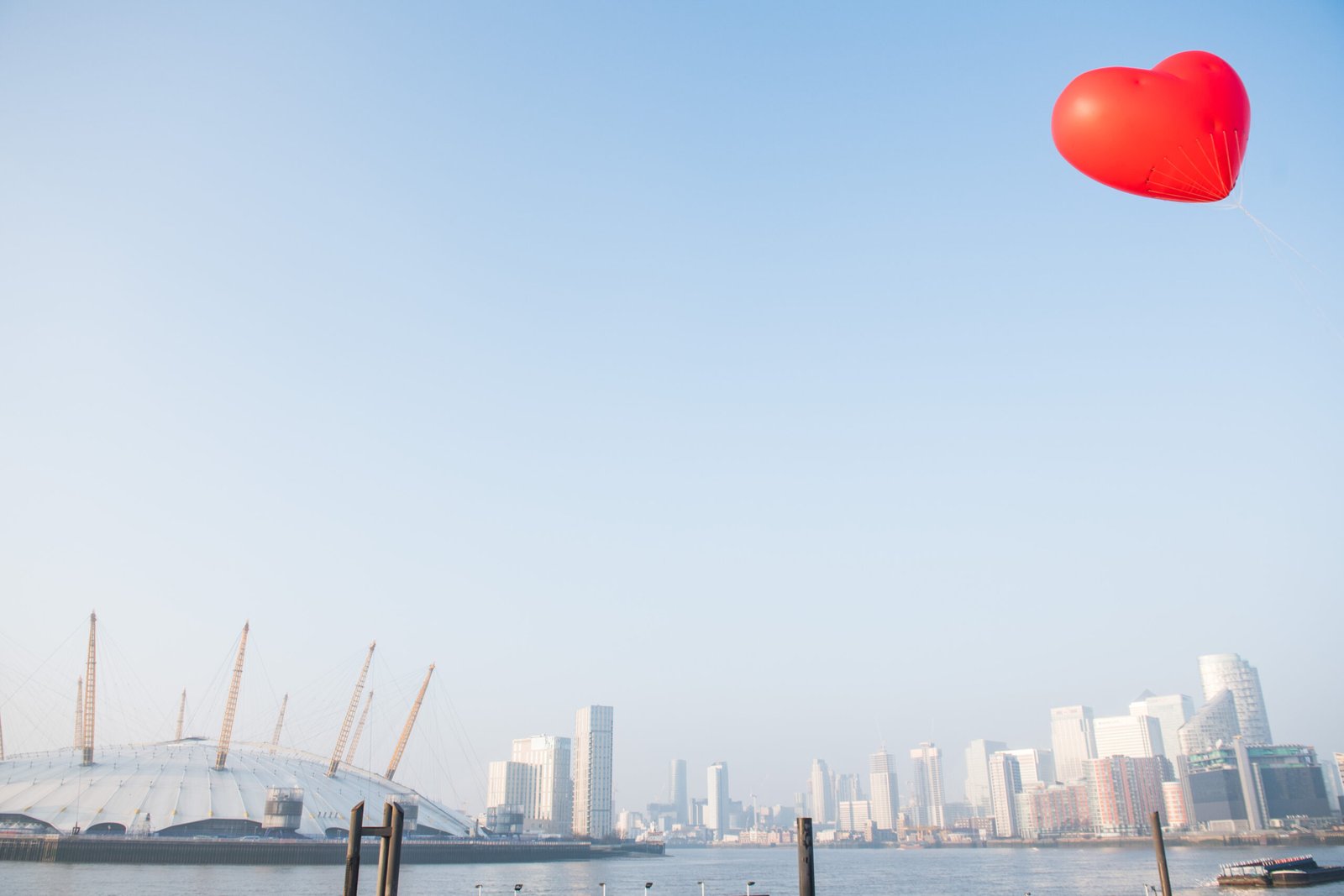 A vibrant red heart-shaped balloon floats in a clear blue sky above a cityscape, captured brilliantly by a corporate photographer in London. Below, modern skyscrapers and a dome structure adorn the waterfront, creating a lively urban backdrop