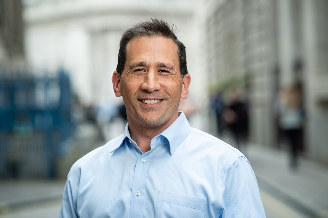 A man in a light blue button-up shirt stands outdoors, smiling at the camera. The background is a city street with blurred buildings and people. James Gifford-Mead Photography - Event Photographer London