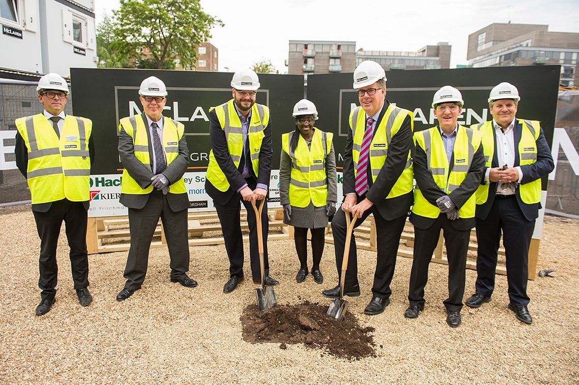 A group of seven people in hard hats and high-visibility vests gather at a groundbreaking ceremony on a gravel area, expertly captured by a corporate photographer. Two individuals hold shovels in the soil, while construction site signs are visible in the background