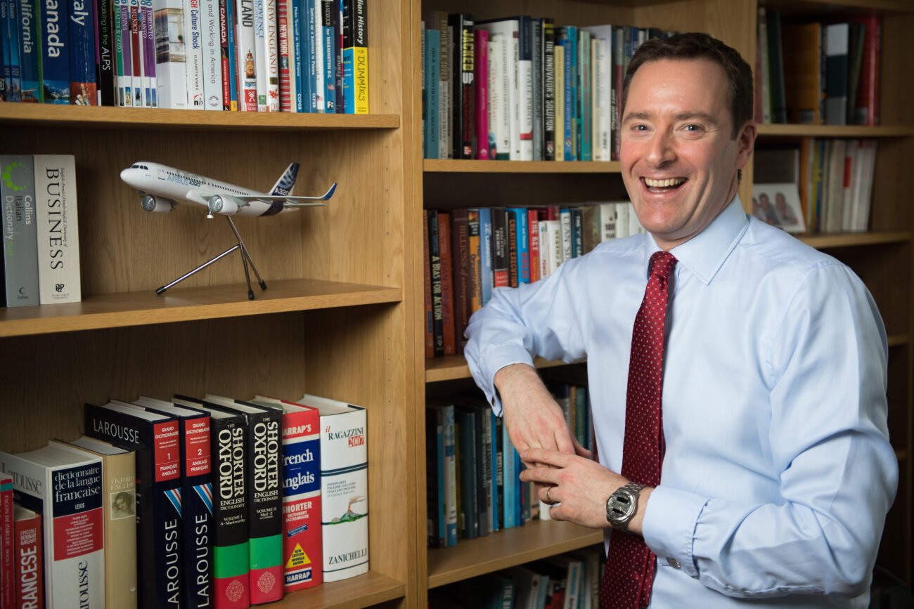 A person in a light blue shirt and red tie is smiling while leaning against a bookshelf filled with books. Captured by a corporate photographer in London, the scene includes a model airplane displayed on one of the shelves, adding flair to this professional setting