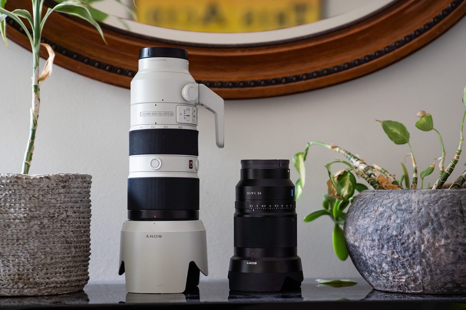 A large white camera lens with a handle and a smaller black lens are displayed on a table between two potted plants. A round wooden-framed mirror is on the wall in the background. James Gifford-Mead Photography - Event Photographer London