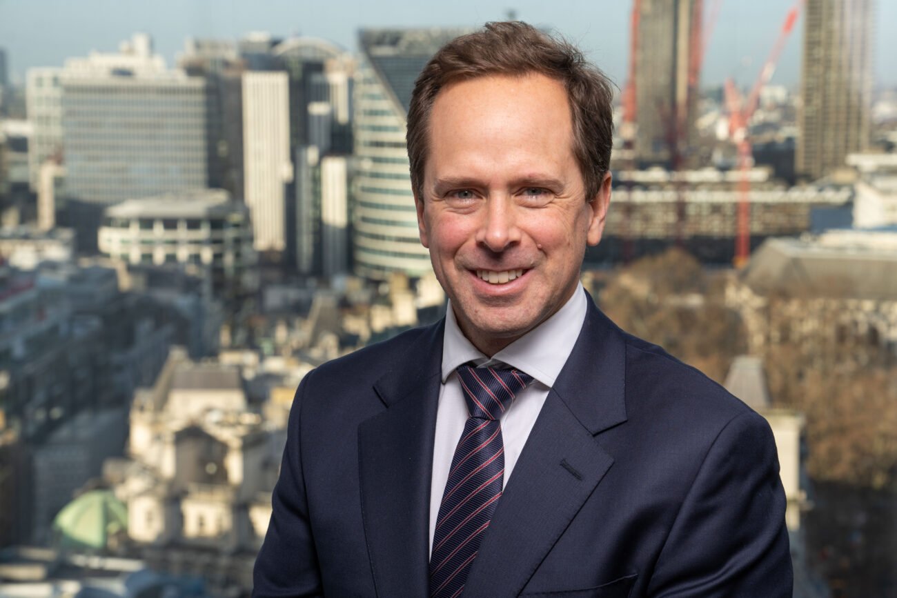 A man in a suit and tie smiles at the camera, embodying the polished style of a corporate headshot. Behind him, the cityscape of tall buildings and cranes basks in sunlight, capturing a perfect blend of professionalism and urban energy. James Gifford-Mead Photography - Event Photographer London