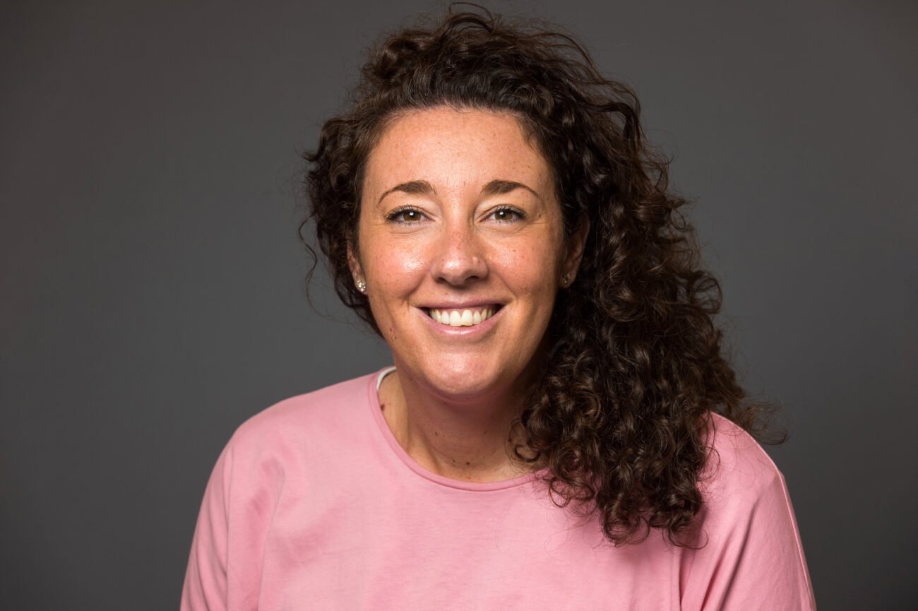 A person with curly brown hair smiles confidently in a pink shirt, poised for their corporate headshot against a plain gray background. James Gifford-Mead Photography - Event Photographer London