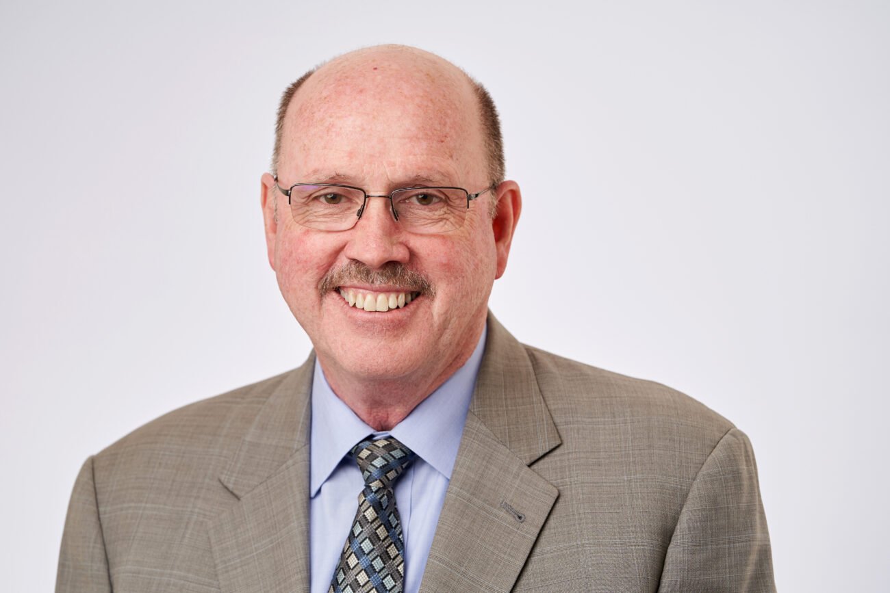 A bald man with glasses and a mustache is smiling for a corporate headshot, dressed in a light gray suit, blue shirt, and patterned tie against a plain white background. James Gifford-Mead Photography - Event Photographer London