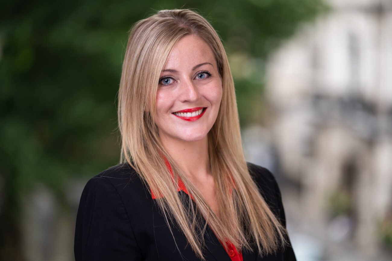 A person with long blonde hair is smiling, wearing a black blazer with a red blouse. Captured by a skilled corporate headshot photographer, the background showcases blurred greenery and buildings, adding an elegant touch to the professional portrait. James Gifford-Mead Photography - Event Photographer London