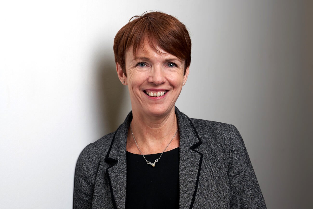 A person with short, red hair smiles at the camera. Captured by a corporate headshot photographer, they exude confidence in a gray blazer over a black top, standing against a plain, light-colored background. James Gifford-Mead Photography - Event Photographer London