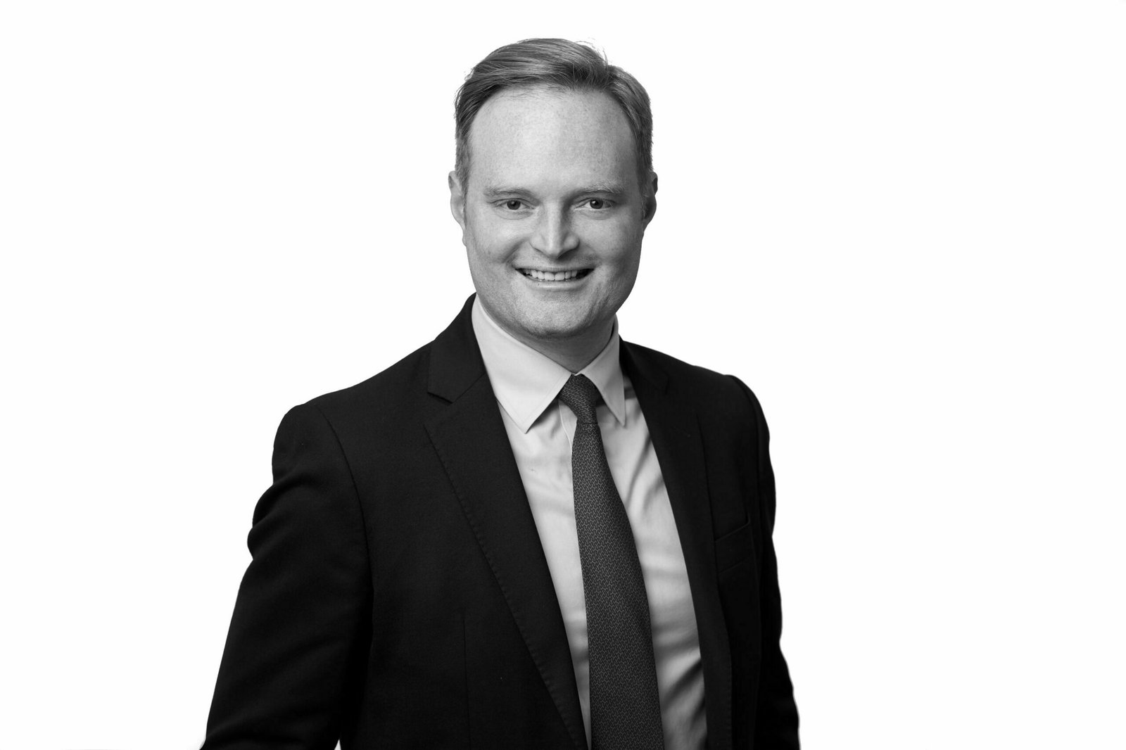 A corporate headshot captures a man in a suit and tie, smiling confidently at the camera against a plain white background. His short hair complements the dark blazer and light-colored shirt, portraying professionalism and approachability