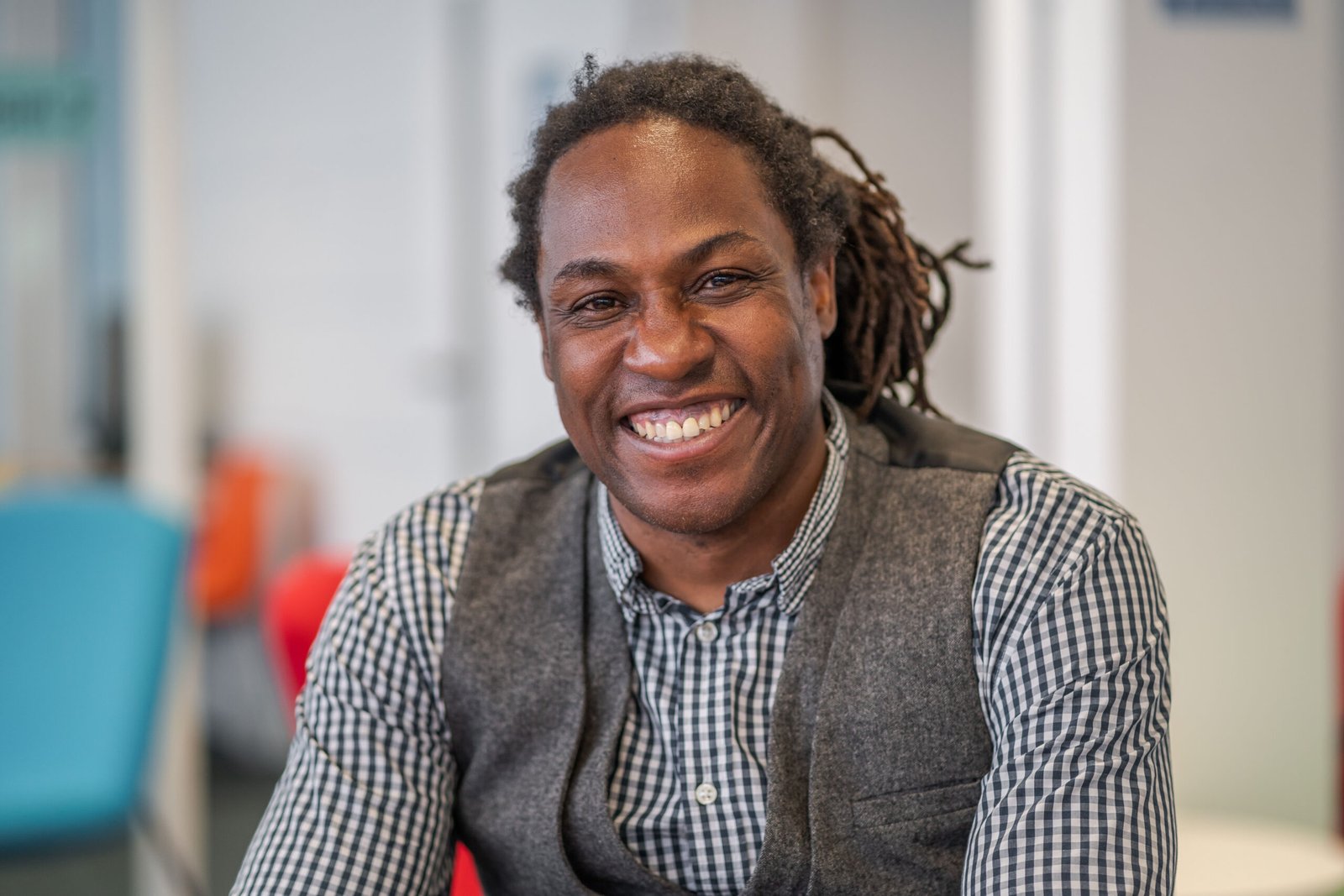 A person smiling warmly, seated in an office captured by a corporate photographer in London. They are wearing a gray checkered shirt with a matching vest. The softly blurred background highlights colorful chairs