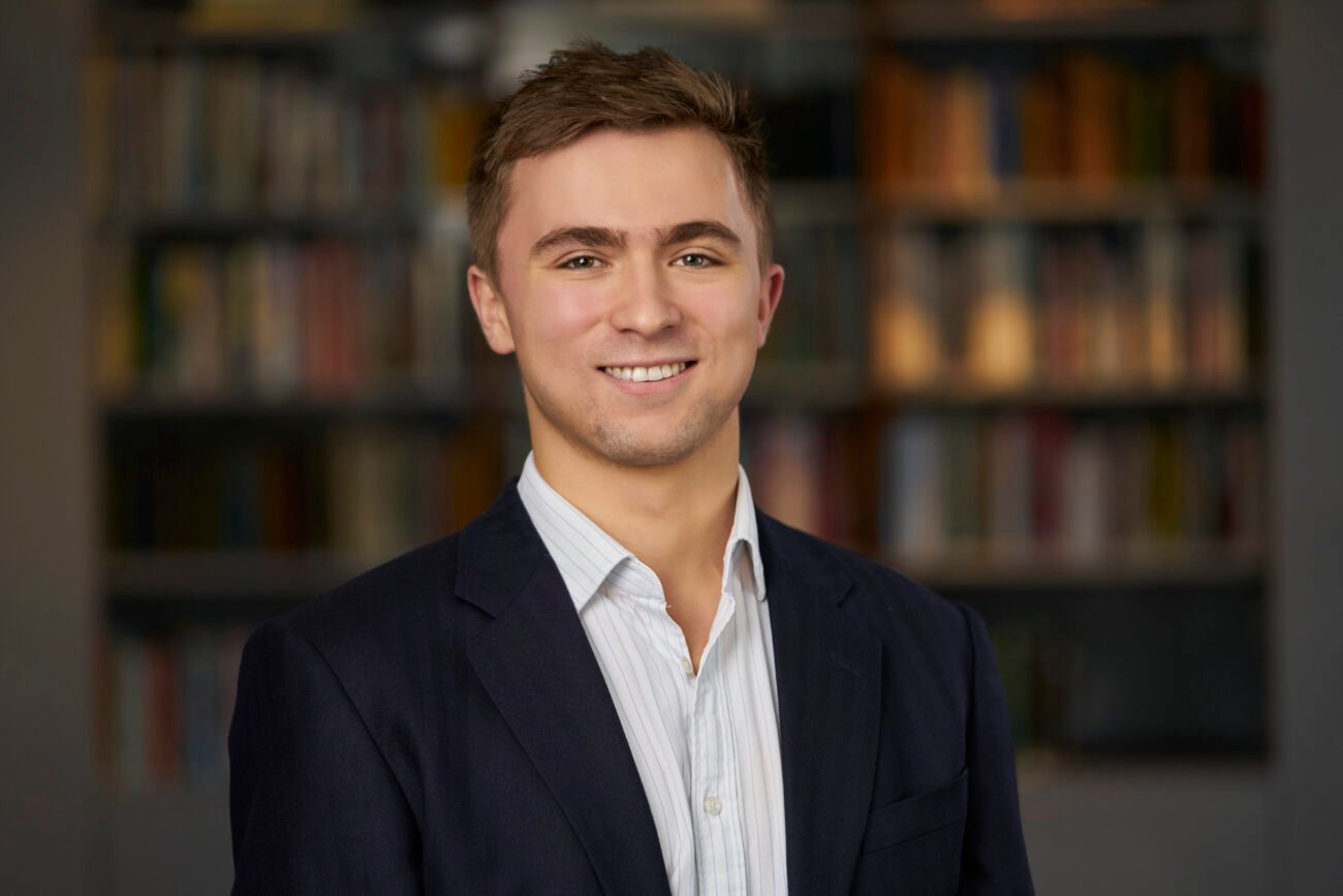 A person in a dark suit and light striped shirt smiles at the camera, captured perfectly by a corporate headshot photographer, with a blurred background of bookshelves adding depth to the image. James Gifford-Mead Photography - Event Photographer London
