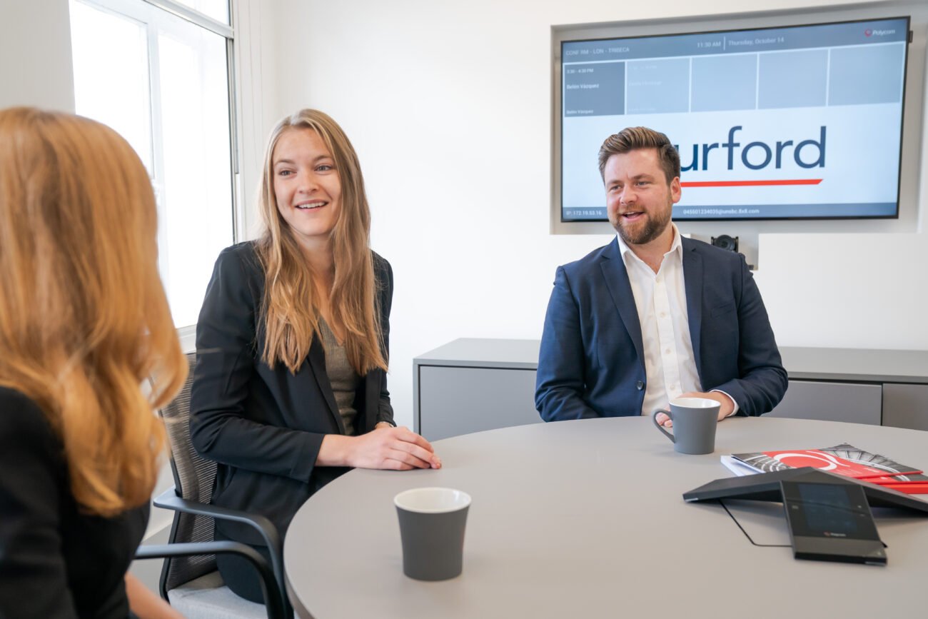 Three people in a business meeting are sitting around a table with coffee cups, captured brilliantly by a corporate photographer from London. A screen in the background displays the word "Burford." They are smiling and wearing business attire