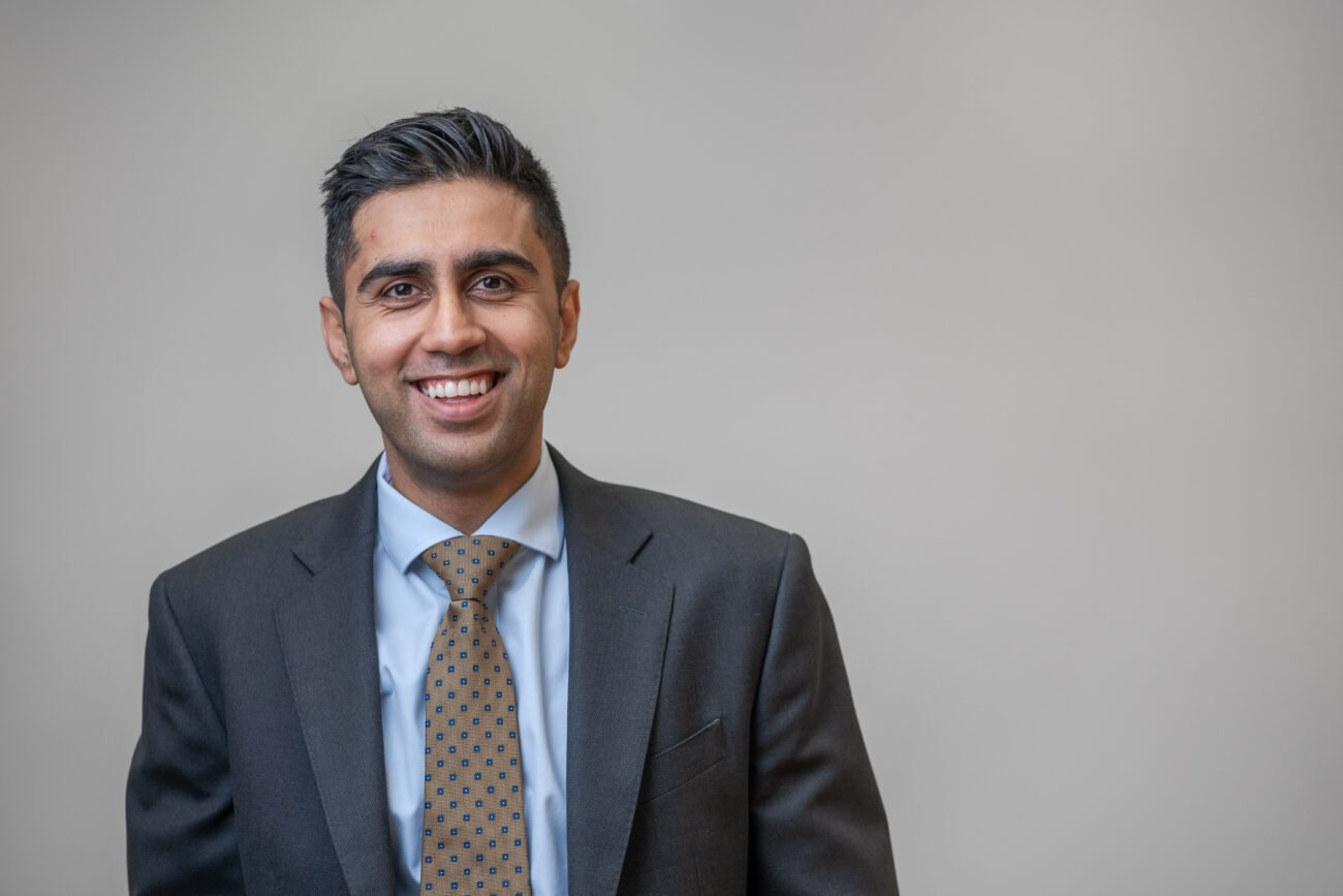 A smiling man in a dark suit with a light blue shirt and patterned brown tie stands against a plain gray background, expertly captured by a corporate headshot photographer. James Gifford-Mead Photography - Event Photographer London