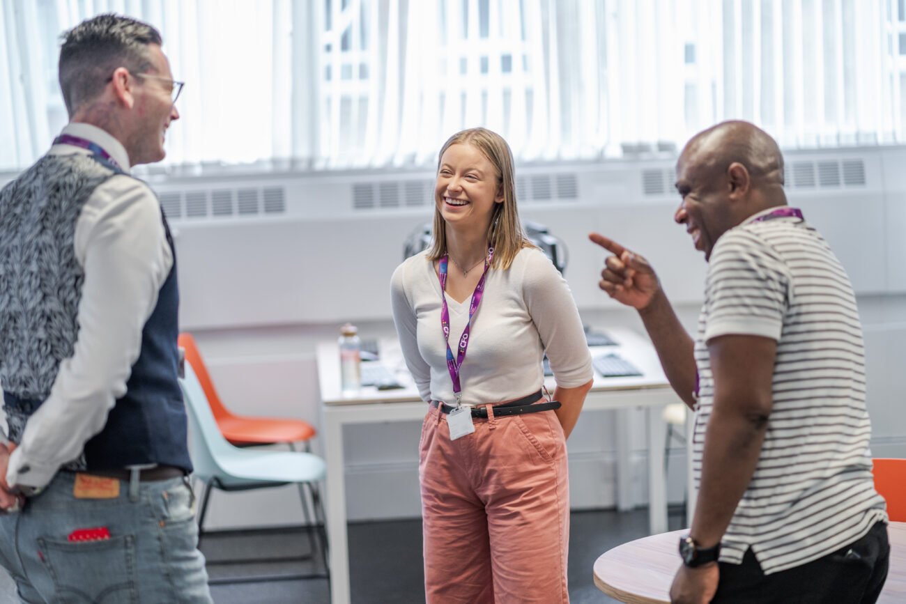 In a brightly lit room, a lively conversation unfolds as three casually dressed individuals wearing ID badges share laughter and smiles. A corporate photographer captures the moment in London, with chairs and a table visible in the background